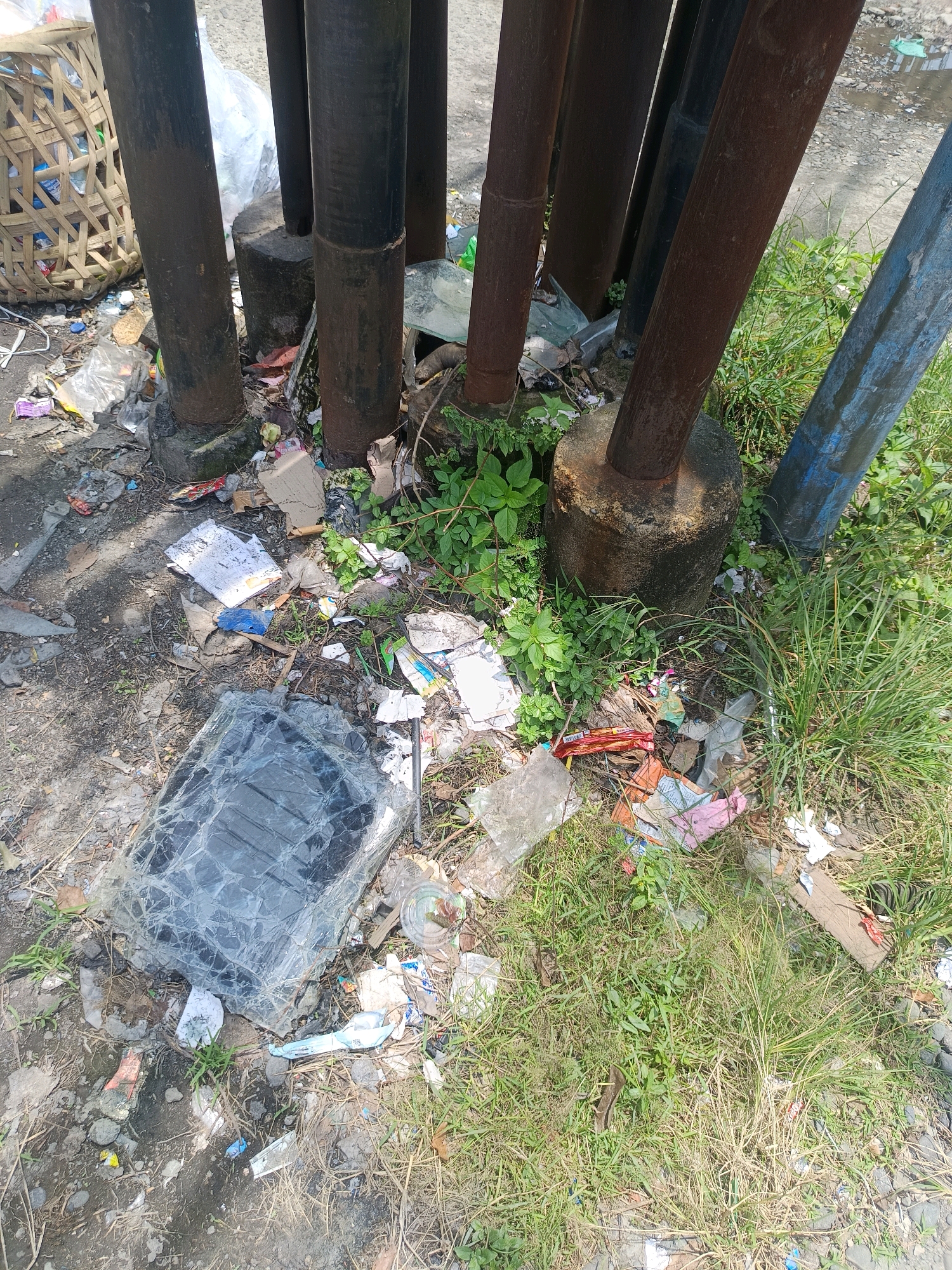 The image shows a scene of litter accumulation around the base of several metal poles, which appear to be structural supports or possibly part of a street light system. The poles are of varying colors, with some rusted brown, one black, and one blue. They are anchored in concrete bases.
The ground around the poles is heavily littered with a variety of refuse. This includes plastic bags, wrappers, pieces of paper, cardboard, and what appears to be a broken piece of dark-colored glass or plastic sheeting. Some green vegetation, including grass and small plants, is growing amongst the debris and around the bases of the poles, suggesting the area may be outdoors and possibly alongside a road or pathway. A woven basket, possibly a trash receptacle, is visible in the upper left of the frame, also filled with some materials. The overall impression is one of neglect and poor waste management. The scene is likely located in an urban or semi-urban environment within Percut Sei Tuan, Indonesia, given the context provided. There are no people or animals visible in the image.