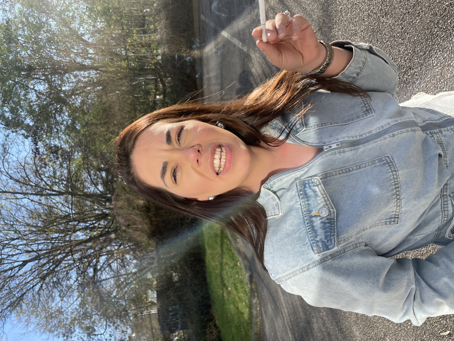The image depicts a female subject outdoors in Montigny-le-Bretonneux, France. She is positioned centrally, capturing her upper body, and is wearing a light blue denim zippered jumpsuit. Her long, dark brown hair flows over her shoulders. The subject's facial expression shows her mouth open, teeth visible, and eyes partially closed, possibly squinting due to the bright sunlight. She holds a lit white cigarette with an orange filter between the thumb and index finger of her right hand, from which smoke is visibly rising in the upper right quadrant. She wears silver-toned stud earrings and a silver metallic watch on her left wrist. The background features an asphalt surface, likely a parking lot or driveway, in the immediate foreground, transitioning to a green grassy area and a dark fence. Behind the fence are numerous deciduous trees, many appearing bare or with minimal foliage, set against a bright blue sky. Strong sunlight, originating from the upper right, illuminates the scene and creates a prominent multicolored lens flare across the left side of the image.