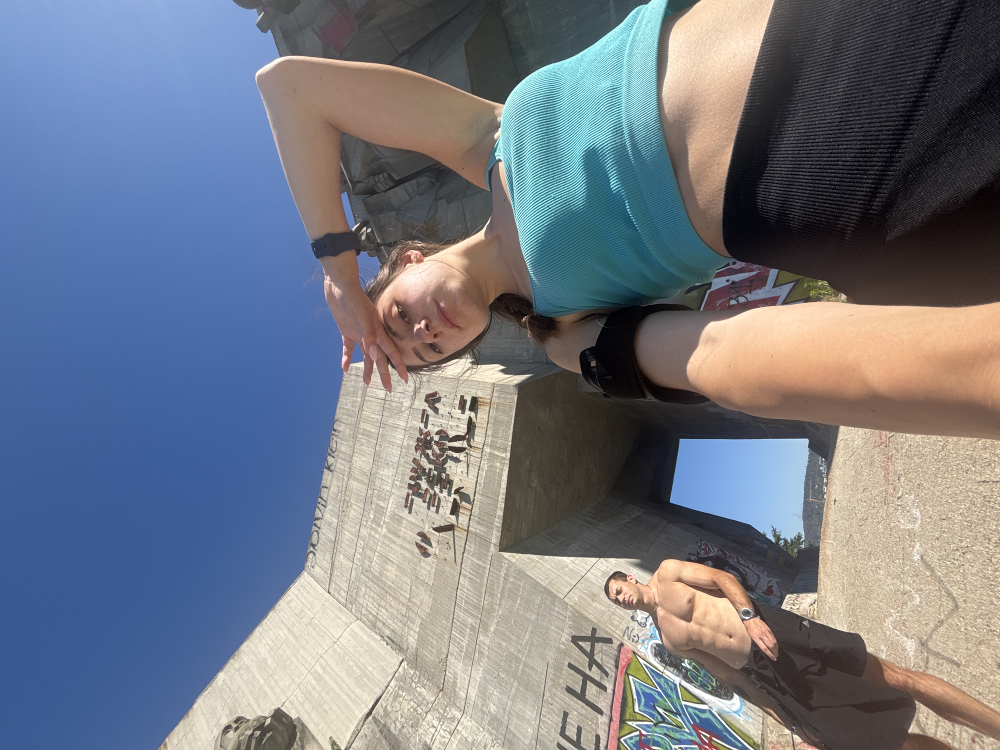 In Varna, Bulgaria, two individuals are captured in a low-angle shot in front of a brutalist concrete structure, possibly a remnant of Soviet-era architecture, heavily adorned with graffiti. The structure features a prominent archway in the background, revealing a clear blue sky.
On the left, a man with a muscular physique, shirtless and wearing black shorts, stands with his arms down. He has a watch on his left wrist. To his right and closer to the camera, a woman is taking a selfie. She is wearing a turquoise ribbed tank top and black athletic shorts. She has an armband on her left bicep. Her right hand is raised to her forehead, shielding her eyes from the sun. The concrete surface they are standing on is rough and shows signs of wear and tear. The overall atmosphere suggests an outdoor recreational activity or exploration.