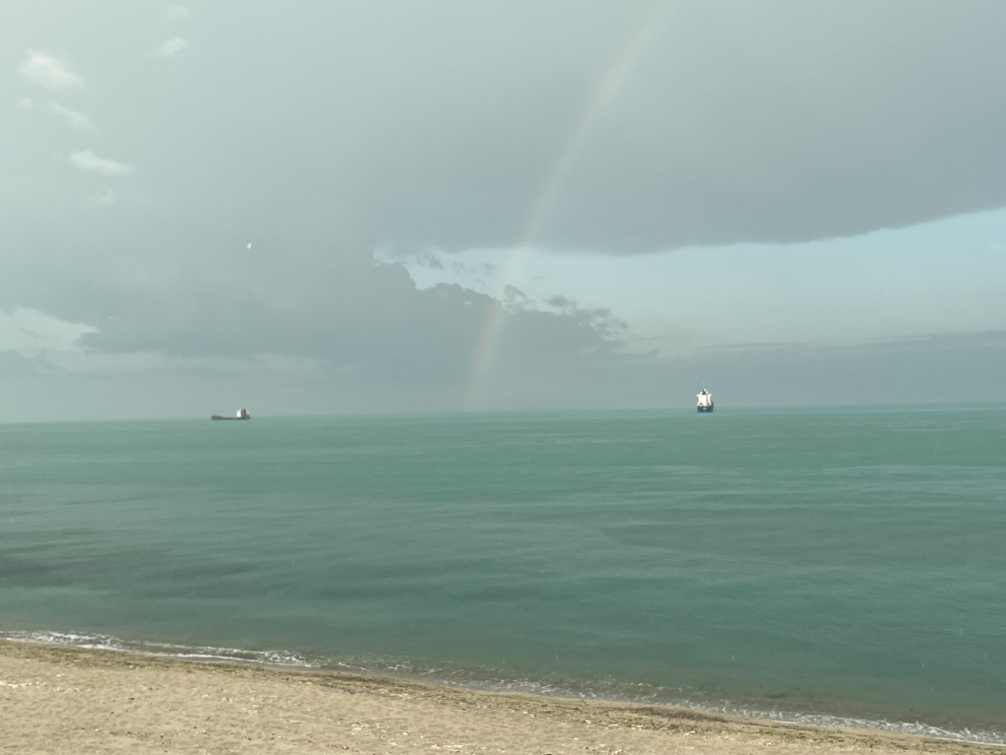 The image shows a coastal scene, likely in Varna, Bulgaria, based on the presence of the Black Sea and common coastal features.
**Environment:** Overcast sky with a prominent rainbow arching across the horizon. The sea is calm, with a greenish-blue hue. A sandy beach is visible in the foreground.
**Objects:** Two cargo ships are visible on the horizon.
**Actions:** There are no actions visible in the image.
**Interactions:** There are no interactions visible in the image.