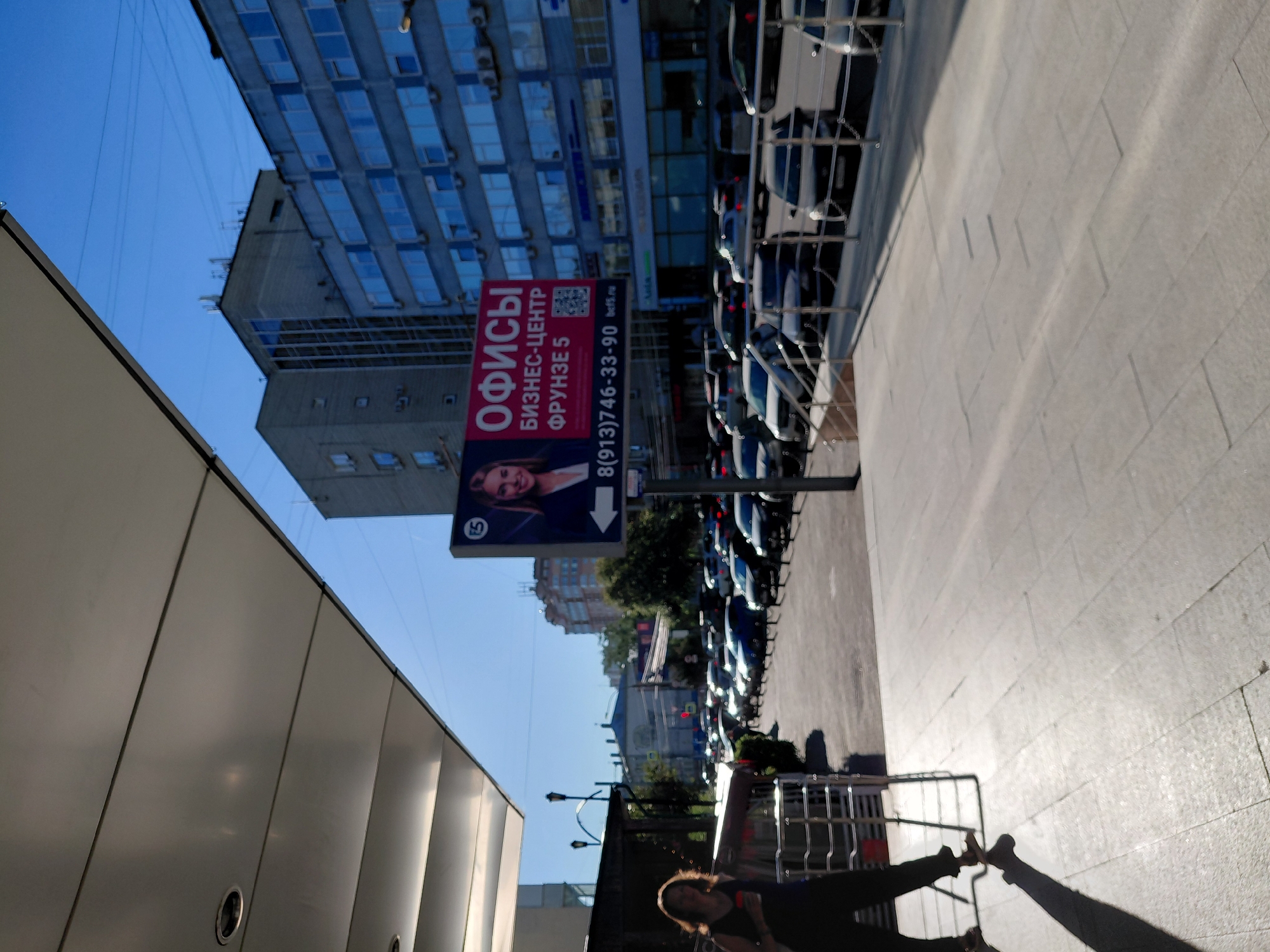 A woman is walking in the foreground on a sunny day in a city. She is wearing a dark top and dark pants, and she is carrying a red cup in her right hand. The background shows a street lined with cars, and beyond that, several multi-story buildings. A large billboard with an image of a woman and text in Russian is visible on the right side of the image. The billboard advertises offices and a business center located on Frunze Street 5. The overall scene suggests a typical urban environment in Novosibirsk, Russia, with clear blue skies and bright sunlight casting long shadows.