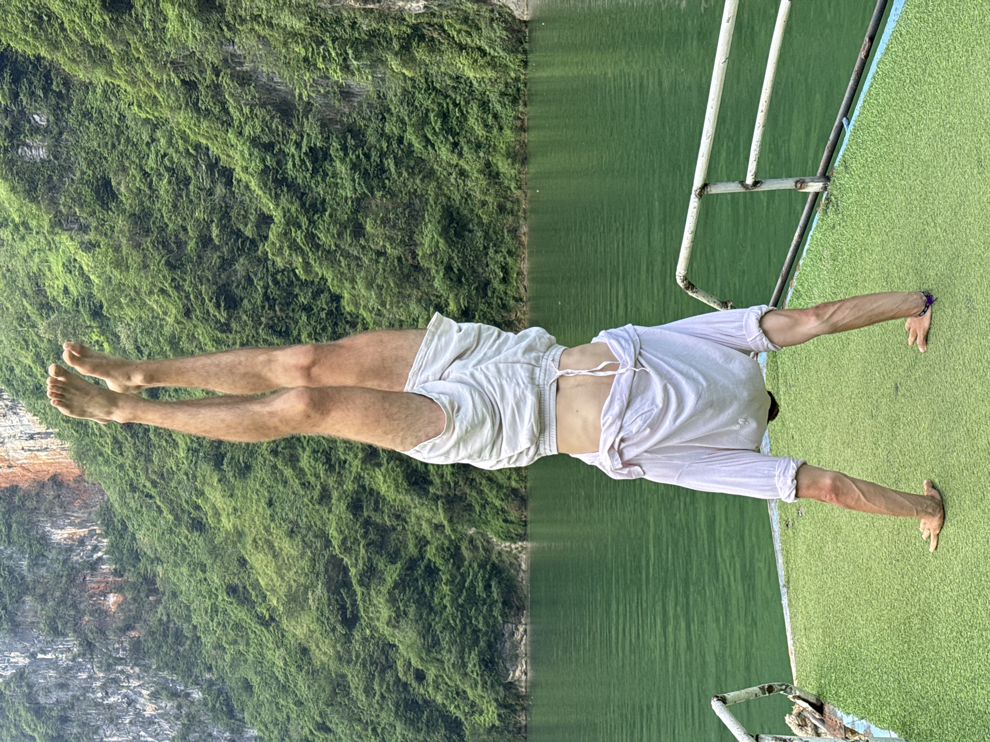 A person in white shorts and a white shirt is performing a handstand on a boat. The boat has green astroturf and a white metal railing. Behind the person, the backdrop is split; on the left is a steep cliff covered in dense green vegetation, and on the right is the still, green water of a river or lake. The location is Đồng Văn Commune, Vietnam, based on the landscape.