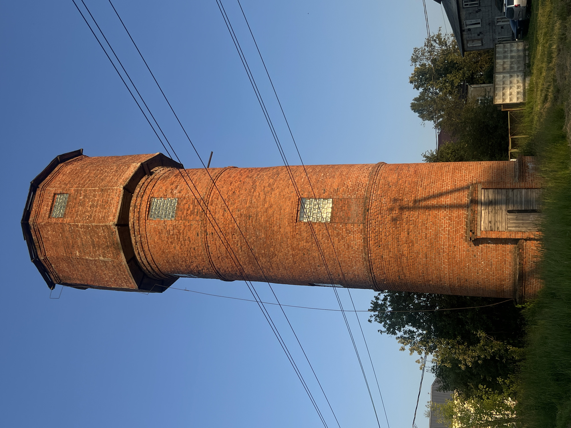 The image shows a tall, cylindrical brick water tower with a hexagonal top, situated in the village of Mostovik, Russia. The tower is constructed of reddish-orange brick and has several small, square windows with translucent glass panes visible along its height. A wooden door is present at the base of the tower. Overhead, multiple electrical wires traverse the frame. Surrounding the tower is a grassy area with trees and other buildings in the background, including what appears to be a residential building with grey siding and a fence. The sky is clear and blue, suggesting a daytime setting.