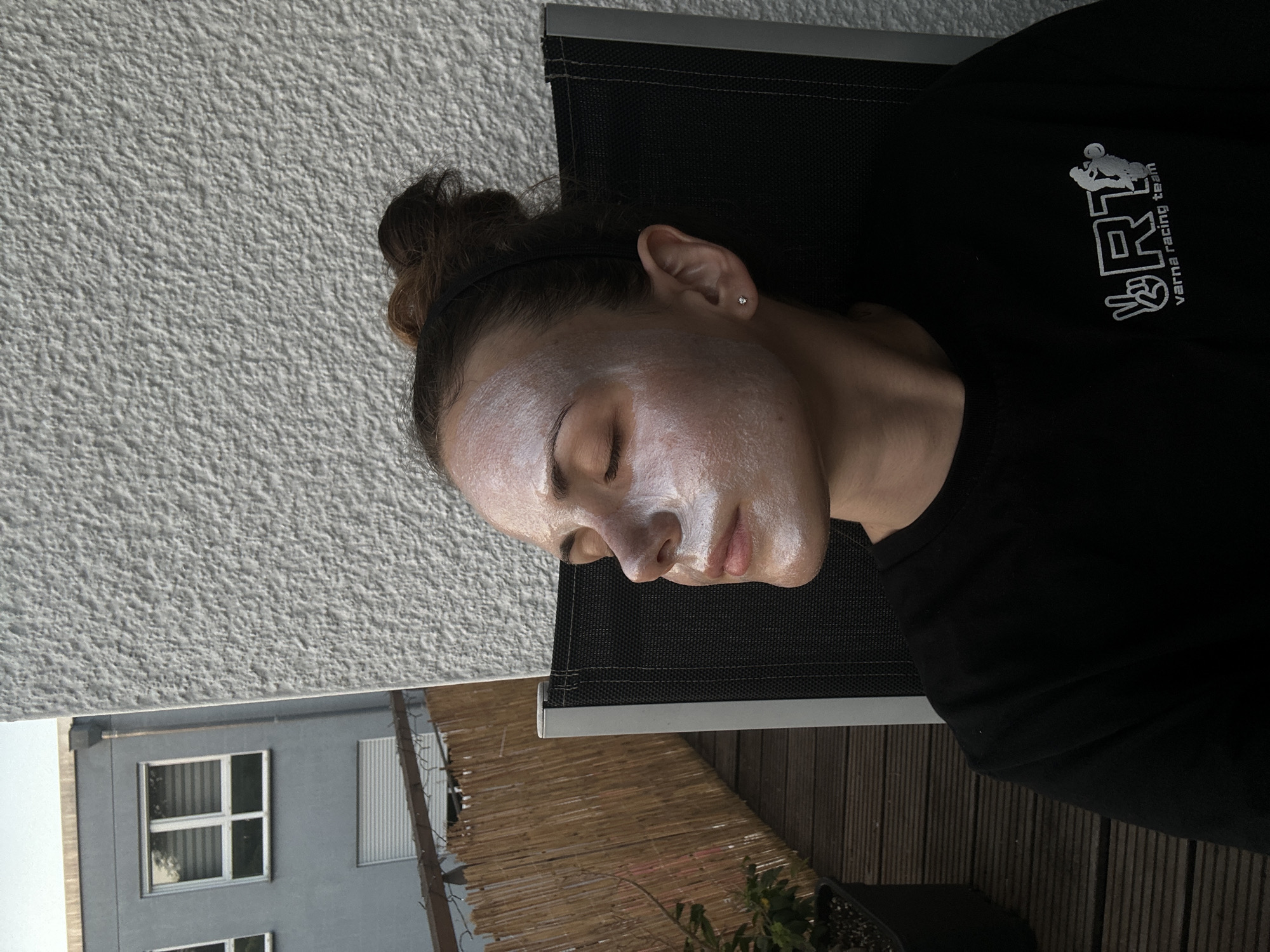 A woman with fair skin and a white facial mask is lying on a black lounge chair, wearing a black t-shirt with a light-colored "Varna Racing Team" logo on the right side of the frame. Her eyes are closed and her hair is pulled back in a bun. The background includes a textured white wall, part of a gray building with windows, and a wooden deck. Some foliage is visible at the bottom. The location appears to be outdoors, possibly on a balcony or patio in an urban setting. The specific city and location are unknown.