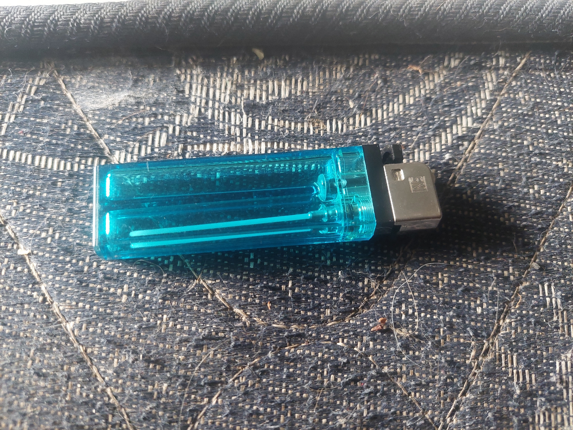 A blue plastic lighter with a silver metal base rests on a dark, textured surface with visible fibers and debris. The lighter is positioned diagonally. The surface appears to be fabric, possibly upholstery or denim, with a woven pattern. The background is out of focus, suggesting a shallow depth of field. The image was taken indoors, likely in a residential setting in Dramaga, Indonesia, given the context provided. There are no people or animals visible in the image, and no discernible actions are taking place. The lighting suggests an indoor environment, possibly with natural light contributing to the scene.