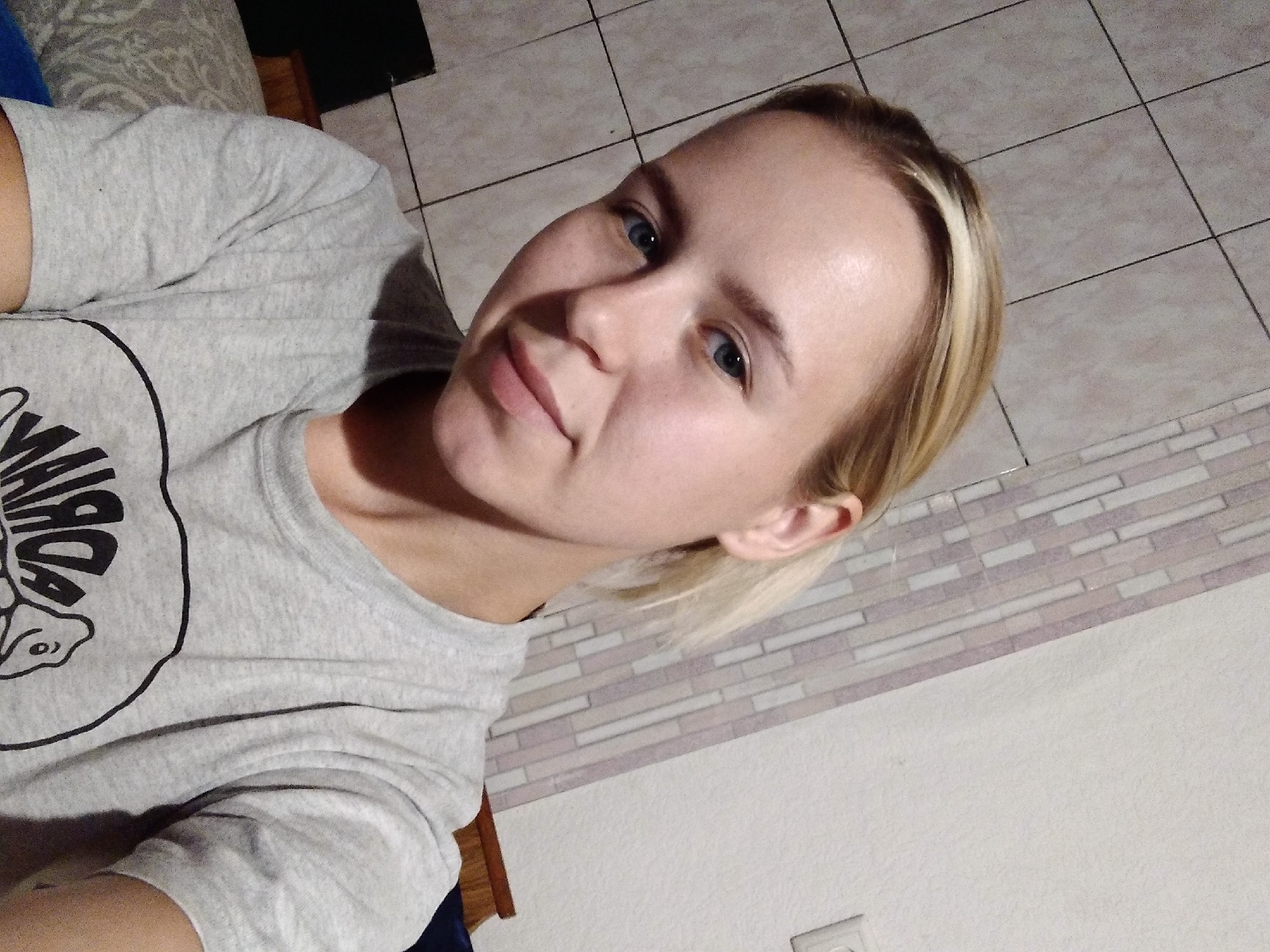 A young woman with blonde hair and blue eyes is taking a selfie. She is wearing a grey t-shirt with a design on it. The background appears to be a wall with tiled sections, suggesting an indoor environment, possibly in Radisne, Ukraine.