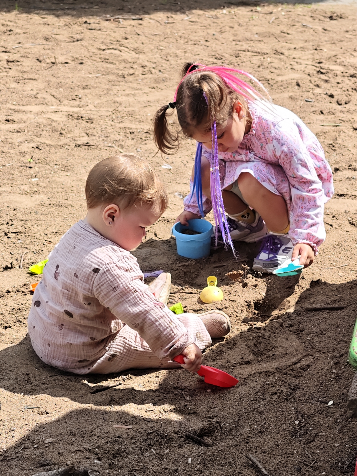 Two young children are playing in a sandpit, likely in a park or backyard. A baby is seated on the sand in the foreground, wearing a light pink, patterned onesie and a pink shoe. The baby is facing to the left, looking down at the sand, and holding a red plastic shovel. To the right and slightly behind the baby, a young girl is crouched down. She has dark hair with pink and purple hair extensions, and she is wearing a pink patterned dress over shorts and sneakers. The girl is looking down into a blue plastic bucket filled with sand. The sandpit is filled with loose, dry sand, with some small twigs and debris scattered on the surface. The lighting suggests it is daytime, with shadows cast on the sand. The image does not provide any specific visual cues that can be used to determine the location within the city of Chaikovsky, Russia.