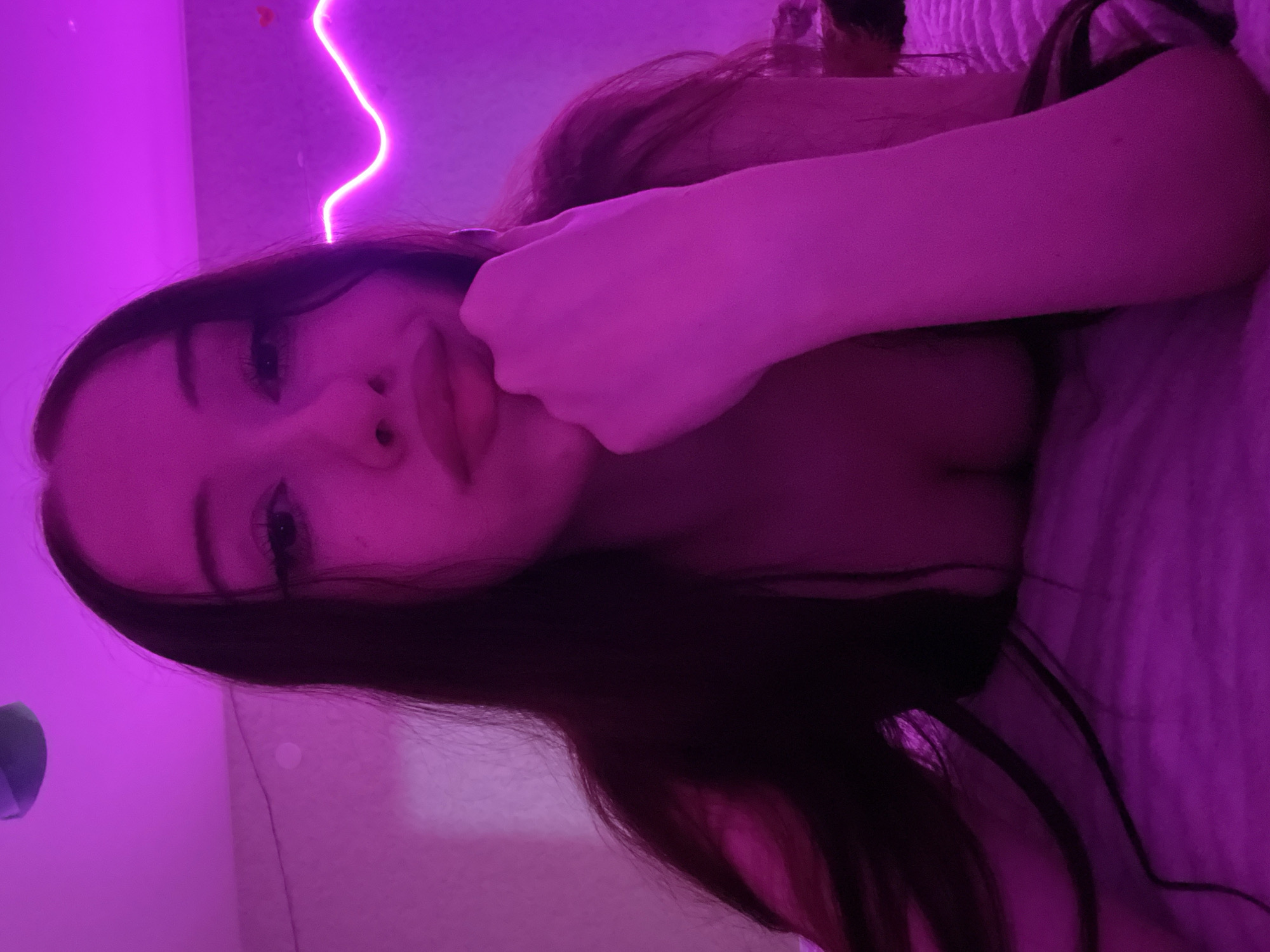 A young woman with long, dark hair is lying down, her face illuminated by purple light. She rests her chin on her hand, her eyes looking towards the camera. Her lips are slightly parted. The background features a pink neon light strip shaped like a wavy line, and a white wall with some slight texture. The overall lighting is a vibrant pink-purple hue. The image does not provide any contextual information to determine its location in Votkinsk, Russia, or any other city.