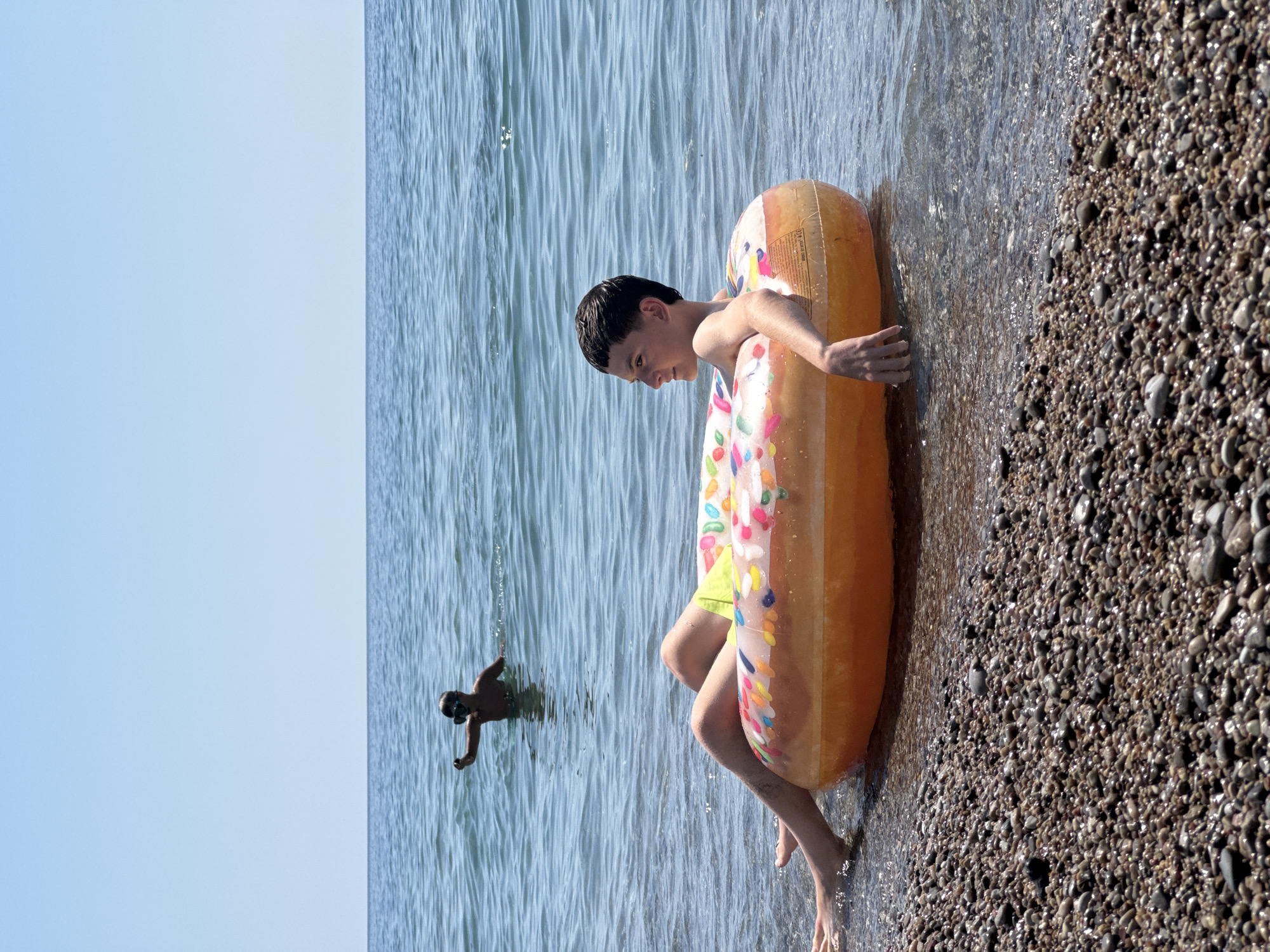 In Đurovići, Montenegro, on a sunny day with clear blue skies, a young boy is floating in the sea on a donut-shaped inflatable. He is lying on his back with his legs extended and resting on the inflatable, which is partially submerged in the shallow, clear water. His arms are relaxed, with one hand resting on the edge of the inflatable and the other slightly submerged. He is wearing yellow swim trunks. In the background, another boy, wearing a snorkel mask, is standing in the water and raising one hand. The foreground is dominated by a pebble beach, with small, wet stones visible where the water meets the shore. The sea extends to the horizon, meeting the sky.