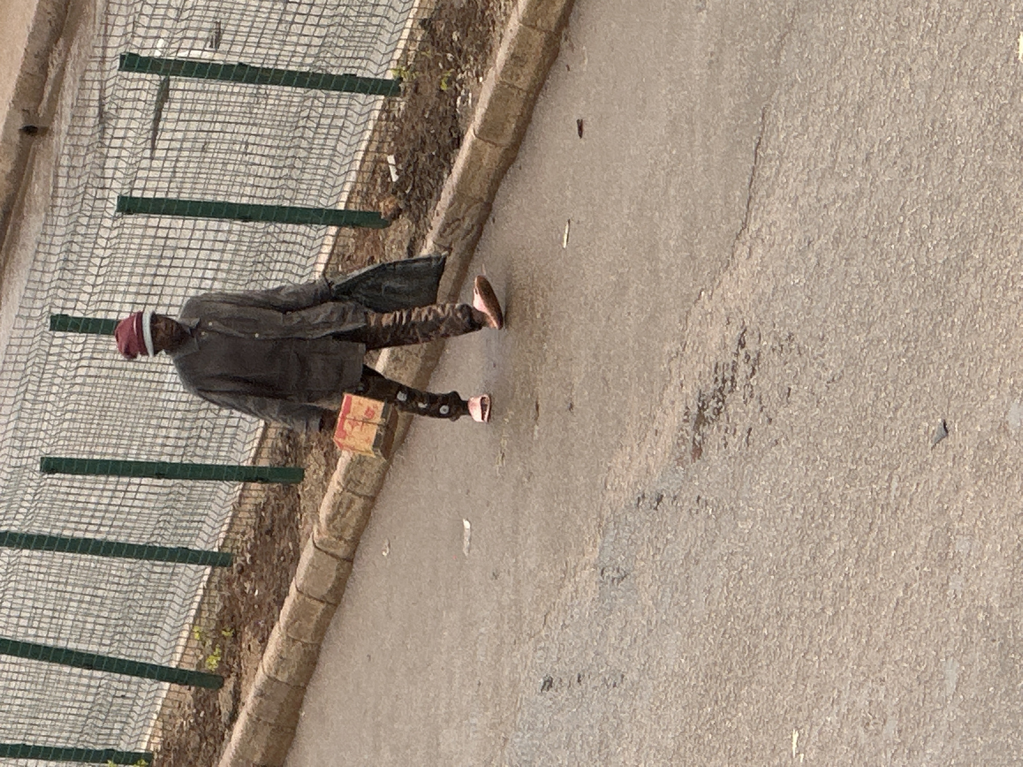 In Bukuru, Nigeria, a man walks along a paved road, his right hand carrying a red and yellow box. He is dressed in a gray overcoat, patterned pants, and a red hat with a white band. In his left hand, he carries a black plastic bag. He is walking near a chain-link fence with green posts, which separates the road from a dirt area with sparse vegetation. The road surface shows signs of wear and some damp patches.