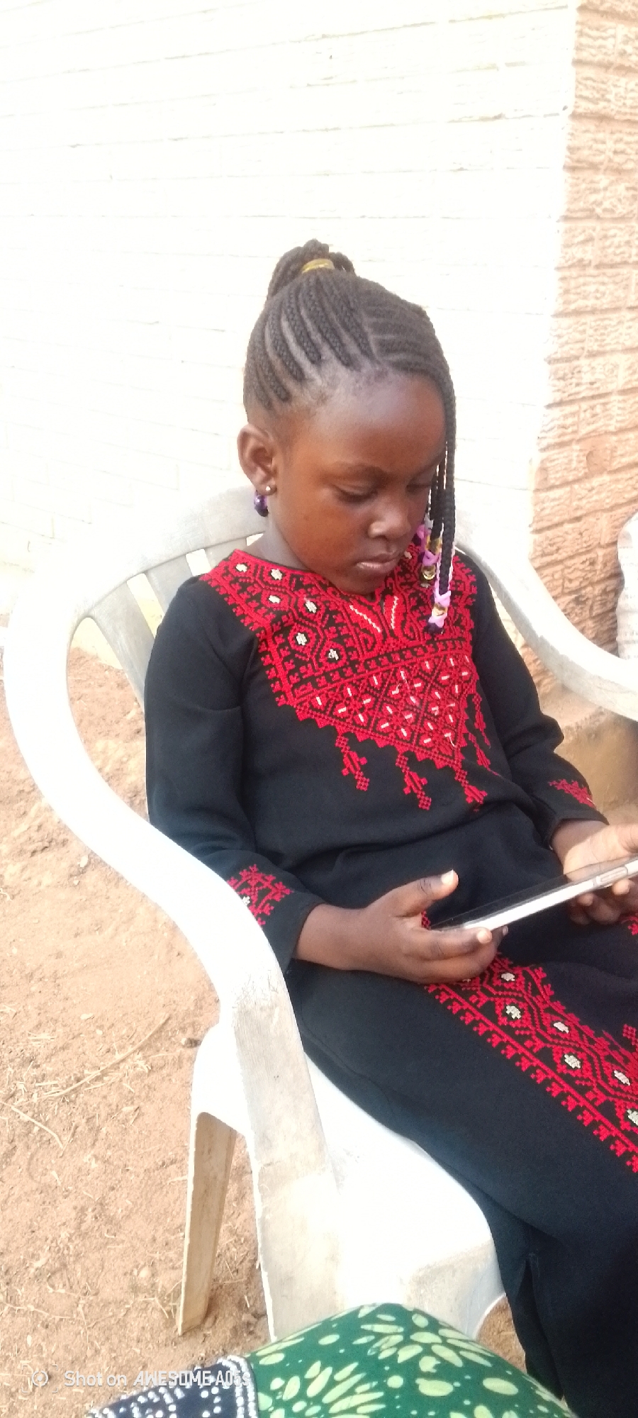 The photo captures a young girl in Narayi, Nigeria, sitting on a white plastic chair, her attention focused on a mobile phone in her hands. She wears a long black dress adorned with intricate red and white embroidery. Her hair is styled in cornrows, some decorated with colorful beads. The backdrop includes a beige wall and a dirt ground. A patterned green and white fabric is also visible.