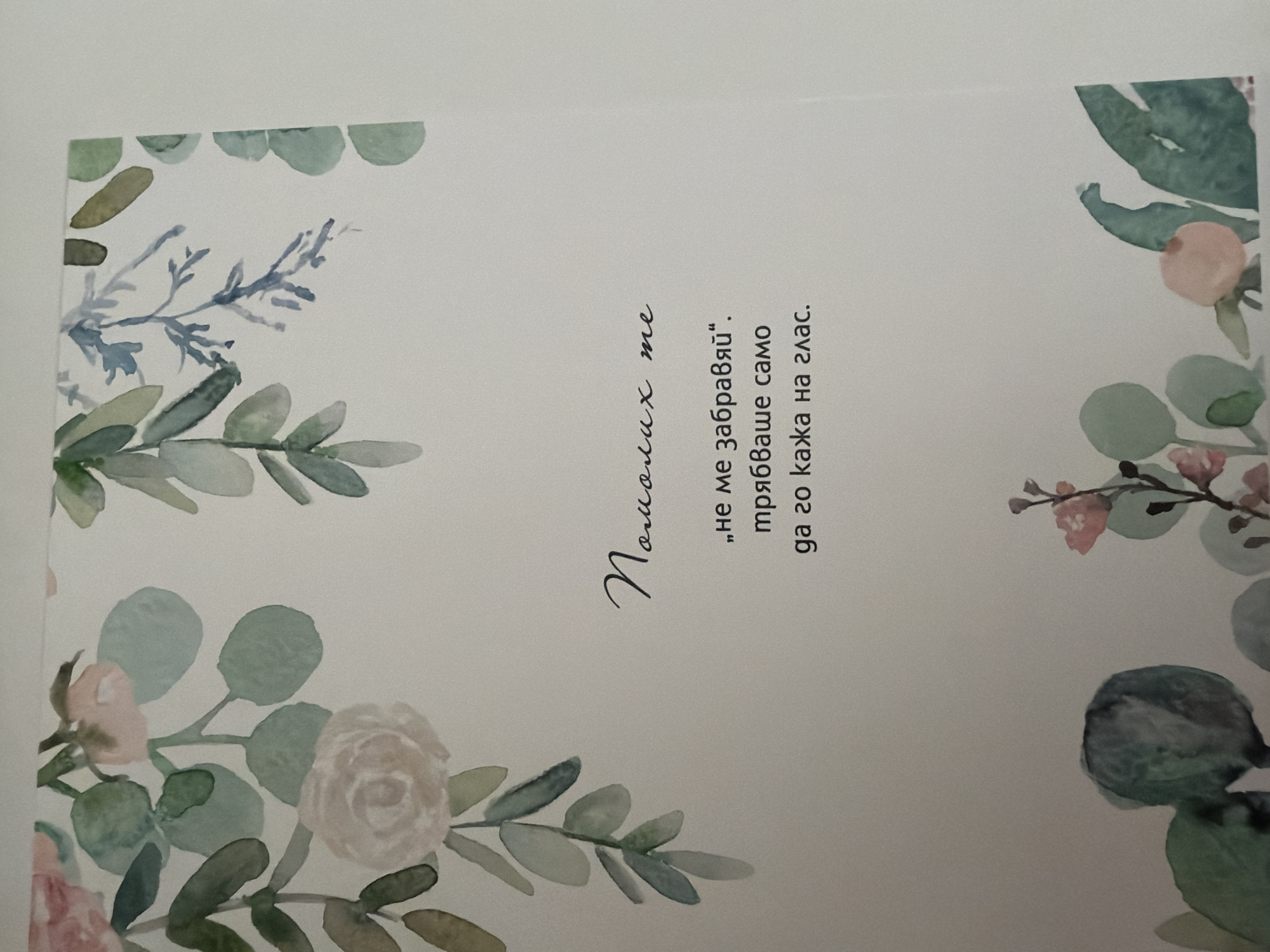 The image is a close-up, slightly angled, overhead shot of a page with watercolor illustrations and text. The page features floral and leaf motifs in muted greens, pinks, and blues framing the text.
The text is in Bulgarian and appears to be a poem or quote. The first line, written in a cursive font, reads "Помних те" (Pomnih te), which translates to "I remembered you." Below this, in a standard serif font, are two lines: "„не ме забравяй“." ("Ne me zabravyay".), meaning "Don't forget me." and "трябваше само" (tryabvashe samo), which translates to "you just had to." The final line reads "да го кажа на глас." (da go kazha na glas.), meaning "say it out loud."
The image does not contain any information that would allow for its location in Pazardzhik, Bulgaria, or any other specific city. There are no people, identifiable landmarks, or contextual clues to suggest a geographic location. The scene is solely focused on the page with the artwork and text.