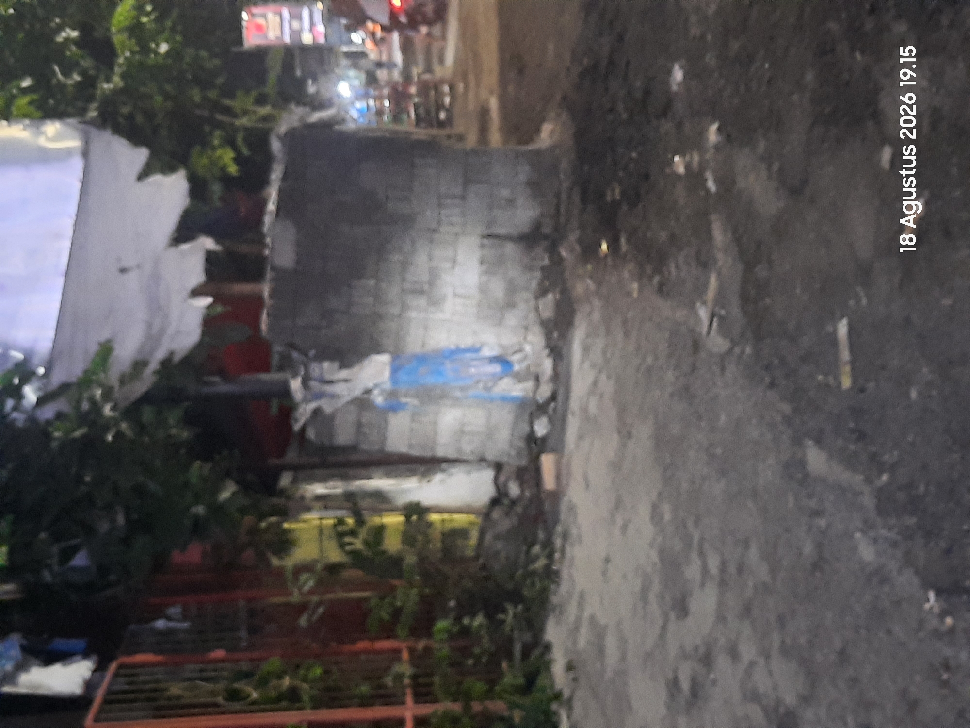The image shows a blurry, low-light outdoor scene, possibly a street or alleyway at night. A rough, gray brick wall occupies the center of the frame, with some blue fabric or material draped over it. To the left, a red metal fence is visible, partially obscured by dark foliage. The ground in the foreground is uneven and appears to be a dirt or asphalt surface with some debris scattered on it. In the background, blurred lights suggest a street or urban environment. A timestamp at the bottom right reads "18 Agustus 2026 19.15". The location is stated as Sukokerto, Indonesia, however, this is not visibly represented in the image. There are no identifiable people or specific activities depicted in the scene.
