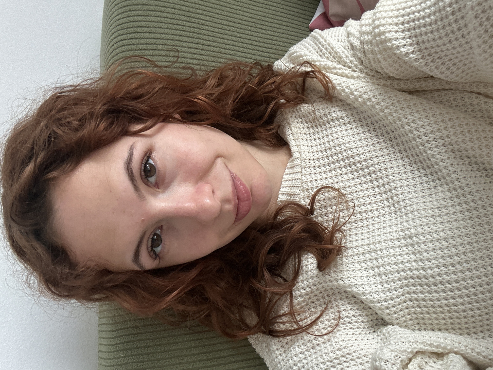 A casual, close-up portrait of a young woman taken indoors, likely a selfie. The image is oriented diagonally, suggesting the woman is lying down. She has reddish-brown, wavy hair that frames her face and spills onto a light-colored, knitted garment. Her brown eyes are open and directed towards the viewer, and she has a soft, slight smile on her lips. Her skin appears clear and natural.
She is wearing a cream or off-white knitted sweater with a textured pattern, which covers her torso and arms. Her hair contrasts with the light color of the sweater. In the background, to the left and top, is a plain white wall. To the right and beneath her head is a textured, ribbed surface in a muted green color, likely a sofa or cushion, providing a soft backdrop. A hint of a light pink object, possibly a pillow, is visible in the upper right corner.
The lighting is soft and natural, indicating daytime, creating a gentle and relaxed atmosphere. The woman appears to be comfortable and at ease, perhaps captured in a moment of rest. There is no visible text in the image. The scene conveys a cozy, domestic setting in Novi Sad, Serbia.
