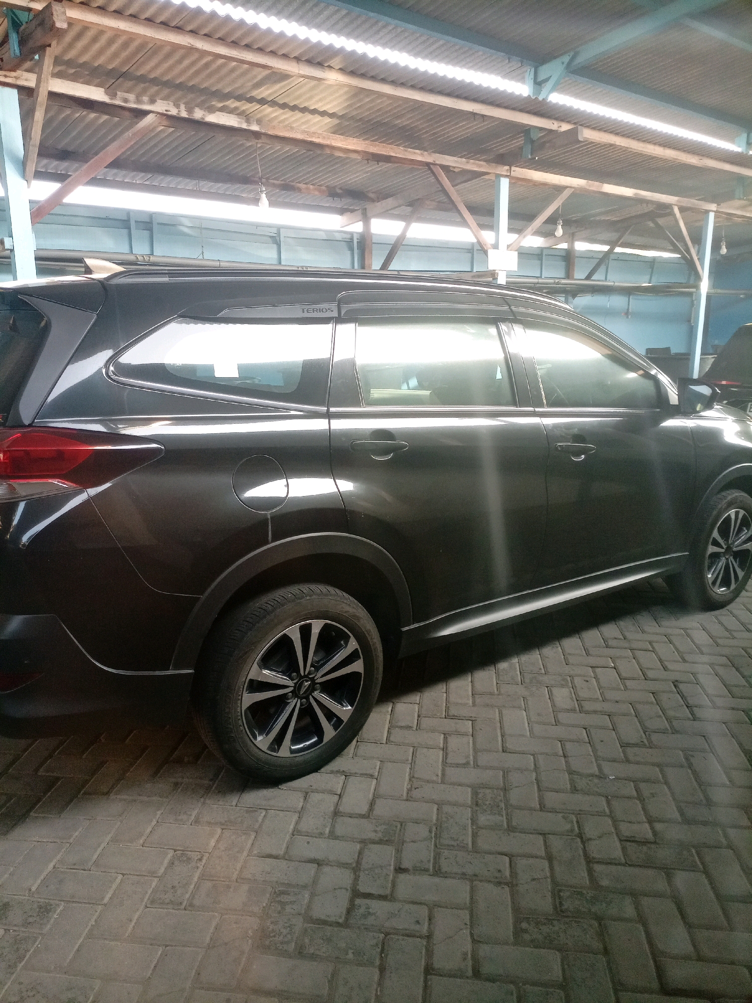 A dark gray SUV is parked on a brick-patterned floor inside a covered structure. The vehicle is viewed from the rear passenger side, showing its profile. The roof of the structure is made of corrugated metal supported by wooden beams. Light filters through gaps in the roofing, casting shadows on the car and the floor. The vehicle has chrome trim around the windows and a silver-colored roof rack. The rear taillight is red and angled. The wheel is a multi-spoke alloy design. The environment appears to be an open-air parking or workshop area, possibly in Sumbersari, Indonesia, suggested by the style of the structure and the brickwork. There are no people visible in the image.