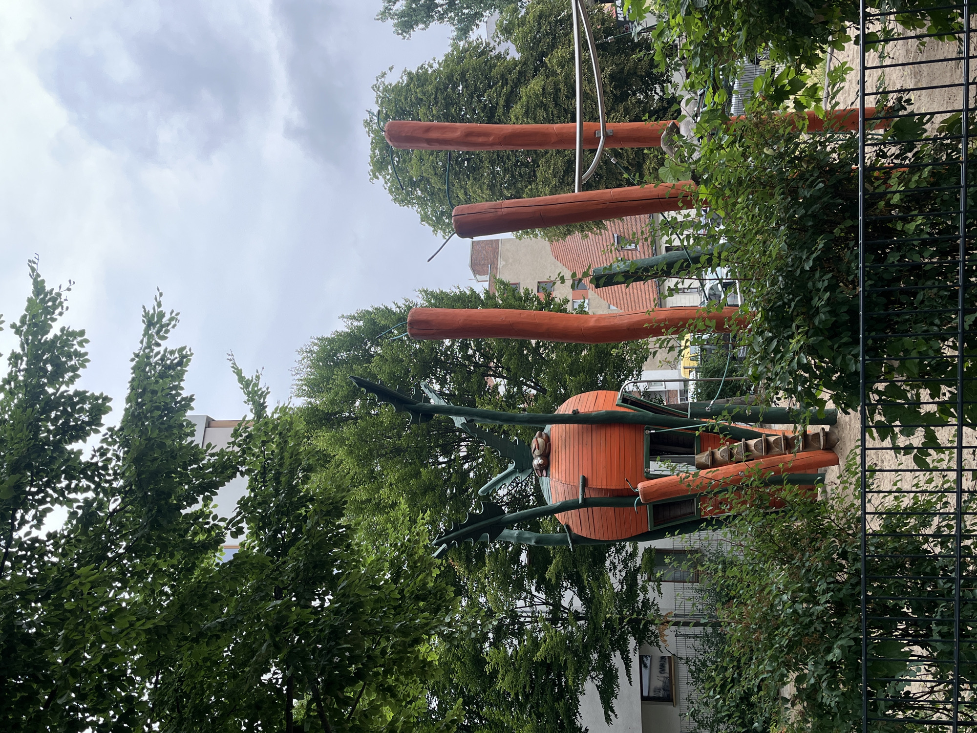 The image depicts an outdoor playground, likely located in a residential area of Berlin, Germany, given the architectural style of the surrounding buildings. In the foreground, a black metal fence encloses a sandy ground. Behind the fence, lush green bushes and small trees are abundant.
The central feature of the playground is a large, orange, spherical structure resembling a carrot, supported by green, twig-like elements. Attached to this structure is a wooden ladder with carved steps. Several tall, orange wooden poles stand vertically around the playground, with some showing green vines growing around them. A silver metal bar is visible connecting some of these poles. In the background, residential buildings and trees are visible under a cloudy sky. The overall impression is that of a whimsical, nature-inspired playground.