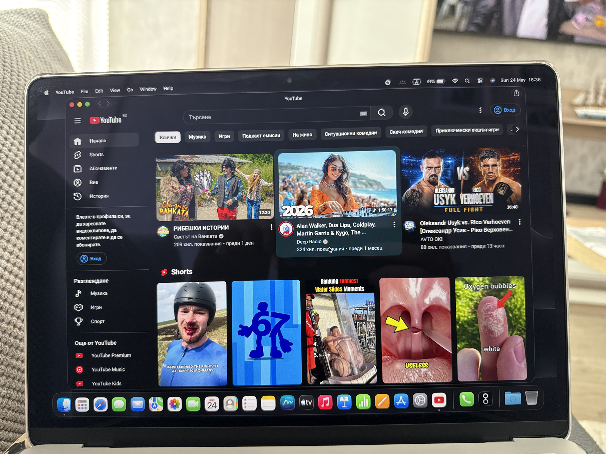 The image shows a laptop displaying the YouTube homepage. The interface is in Bulgarian, with options for "Home," "Shorts," "Subscriptions," and "History" on the left sidebar. The main content area shows video thumbnails, including music videos and boxing highlights. Below, the "Shorts" section features thumbnails with diverse content. The laptop's dock at the bottom shows various application icons. The date and time displayed in the upper right corner of the screen reads "Sun 24 May 16:36." The exact location of Lovech, Bulgaria, is not depicted within the image.