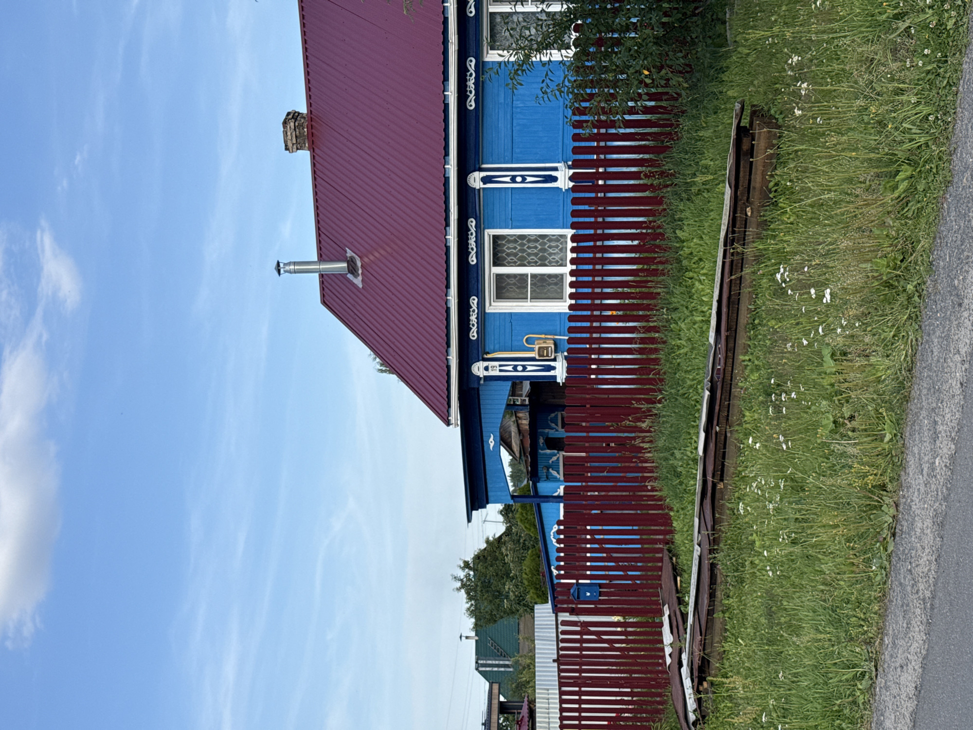 The image depicts a traditional wooden house with a burgundy metal roof. The house is painted bright blue and features ornate white trim around the windows and along the roofline. A metal chimney pipe protrudes from the roof. A dark red wooden fence runs along the side of the house, partially obscuring the view of the building. Beyond the fence, a grassy area with wildflowers grows beside an asphalt road. In the background, other houses with metal roofs and some trees are visible. The sky is a clear blue with some wispy white clouds.
The image appears to be taken on a sunny day, likely in the late morning or early afternoon, given the bright light and shadows. The setting is rural, and the presence of traditional architecture and the natural landscape suggest a location in Russia, possibly a village or dacha settlement. There is no visible activity or people in the scene. The overall impression is one of a peaceful, rural setting.
The visible text is "19" on a white decoration above one of the windows.