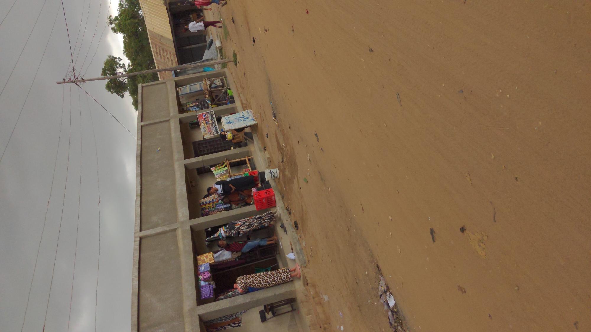 The image depicts a street in a low-income area, possibly in Masaka, Nigeria. There is a dusty dirt road occupying most of the frame's right side. The left side features a partially constructed, multi-story building with several open-fronted stalls. These stalls seem to be used for small-scale trading, with vendors selling items such as fabrics, groceries, and other goods. Some people are visible inside the stalls, apparently conducting business. Overhead power lines are present, and the sky is overcast with dull gray clouds. In the distance, additional buildings and trees can be seen. The general impression is that of a busy, informal commercial area in a developing urban environment.