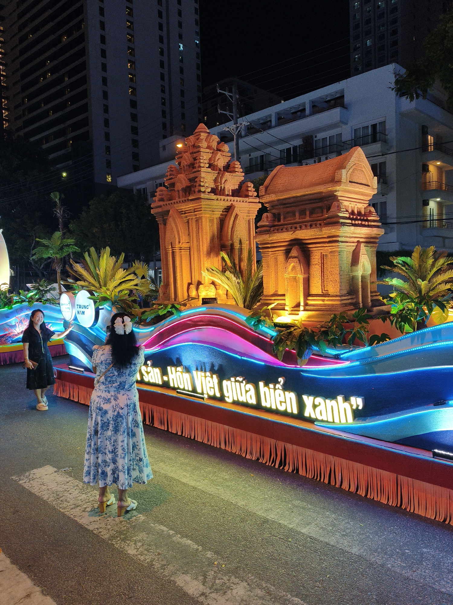 In Nha Trang, Vietnam, at night, a brightly lit parade float is on display. The float features a replica of Cham towers, bathed in warm orange light, surrounded by lush green palm fronds and foliage. The float's base is adorned with colorful neon lights in shades of pink, blue, and purple, mimicking waves. Prominently displayed in large, illuminated white lettering are Vietnamese characters that read: "Sản - Hồn Việt giữa biển xanh" which translates to "Vietnamese Identity amidst the blue sea". Two women are present: one in a black dress stands to the left, looking towards the float and gesturing with her left hand; the other woman, facing away from the camera, wears a blue and white floral dress and is taking a photo of the float with her phone. In the background, tall apartment buildings are visible, their windows lit up, indicating an urban environment. The scene is set on a paved road, suggesting a street during a parade or festival.