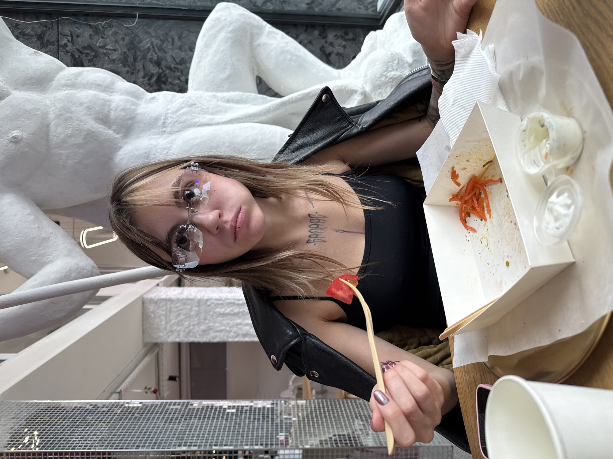 The image captures a young woman with blonde-streaked hair, wearing a black tank top and a black leather jacket draped over her shoulders, sitting at a table indoors. She is holding a small wooden utensil with a piece of tomato on it, poised near her mouth. Her expression is one of mild dissatisfaction or boredom. In front of her is a partially eaten meal in a white takeout container, with some shredded carrots and remnants of sauce visible. There are also two small plastic containers of sauce or dressing on the table, one open and partially consumed. A white paper cup and a pink smartphone are also on the table. The background features a large, white, abstract sculpture that resembles a human torso, and a wall with a mirrored, mosaic-like surface on the left. The lighting suggests an indoor environment with natural light filtering in. The setting appears to be a restaurant or cafe with a modern, artistic ambiance, possibly in Moscow, Russia, given the context.