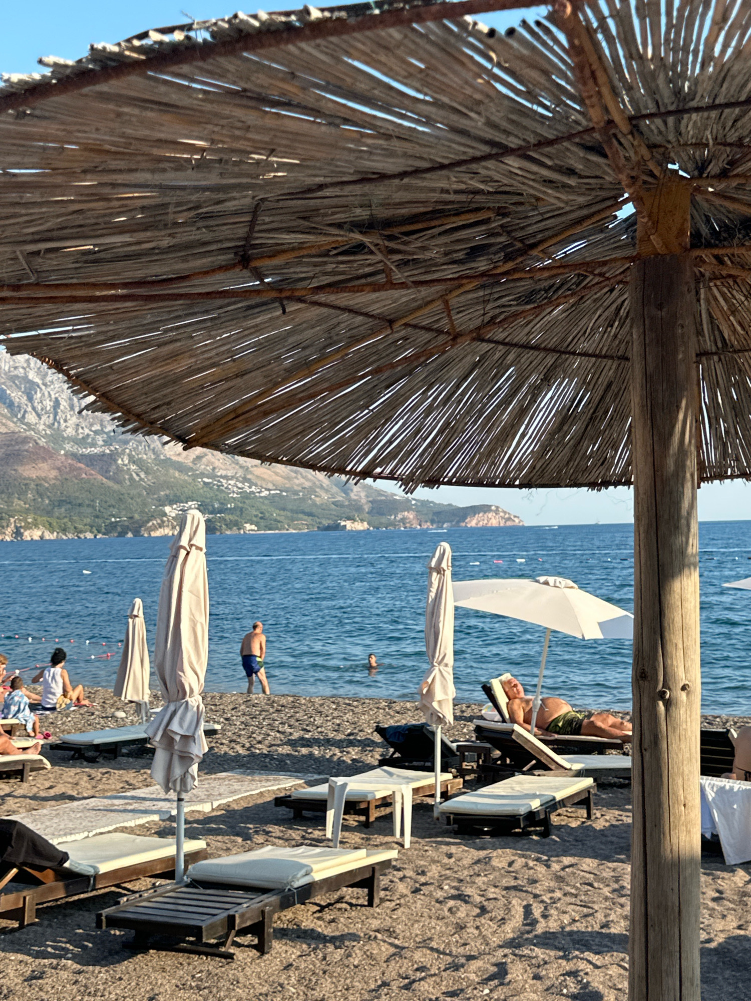 This image captures a beach scene in Boreti, Montenegro. In the foreground, a rustic thatched umbrella with a wooden pole provides shade. Beach chairs and loungers are arranged on the pebble beach. Several closed beach umbrellas stand upright. In the midground, people are visible on the beach and in the blue water. A man in blue shorts is standing in the shallow water, while another person swims further out. A white umbrella shades a person lying on a lounger. The background features the Adriatic Sea with a hilly, mountainous coastline characteristic of the region, dotted with buildings. The sky is clear and blue.