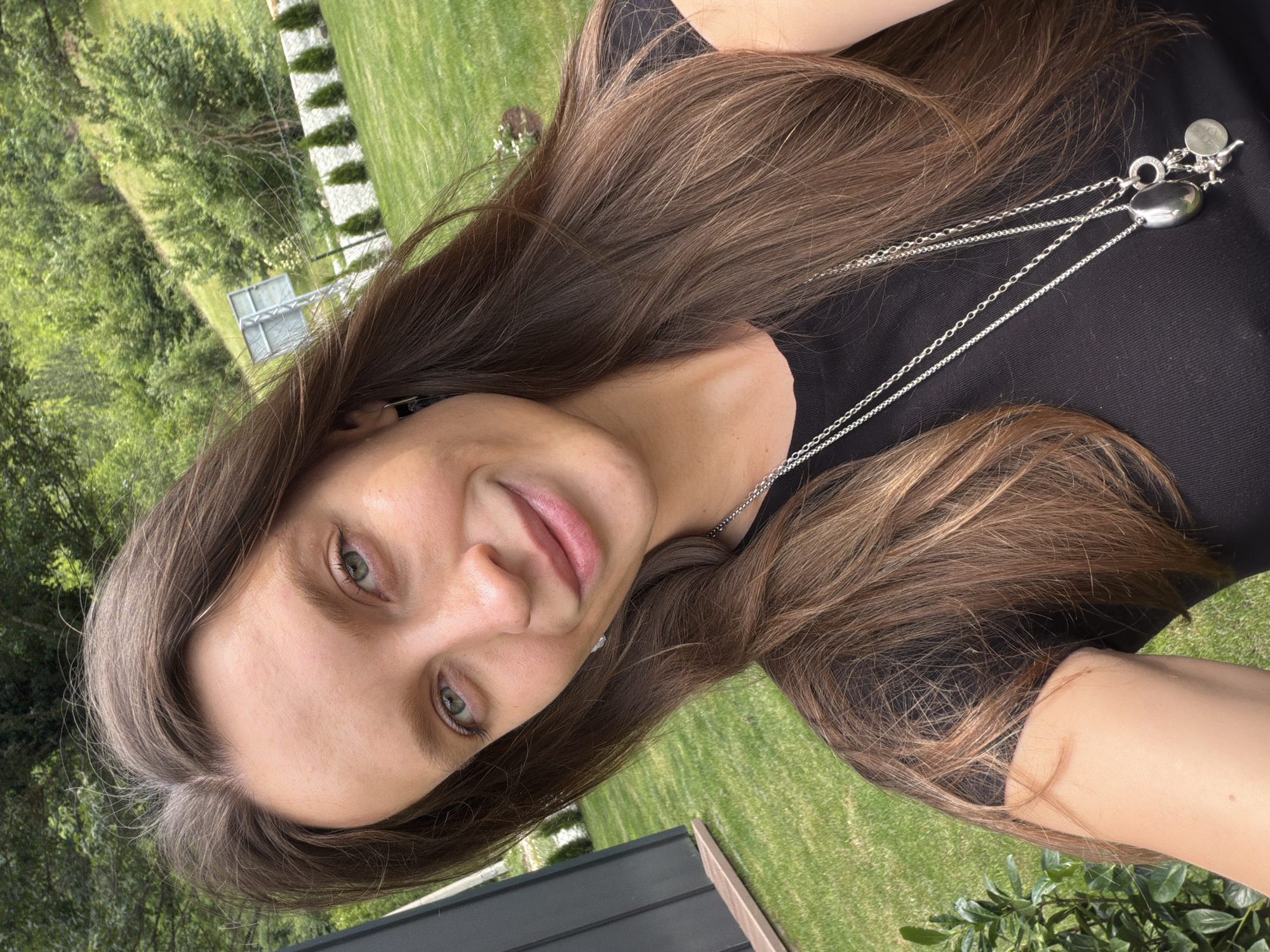 The media file is a portrait of a young woman with long, brown hair, taken outdoors in what appears to be a backyard setting. The woman is wearing a black top and a silver necklace with a pendant. Her face is turned towards the camera, and she has a slight smile. She has green eyes and light skin.
The background consists of a well-maintained green lawn, trees, and some fencing. The lighting suggests it is daytime, possibly late afternoon, with a pleasant outdoor atmosphere. The scene evokes a casual and relaxed mood. There is no visible text in the image. The location context provided is Bistrica, Montenegro, which aligns with the natural outdoor scenery depicted.