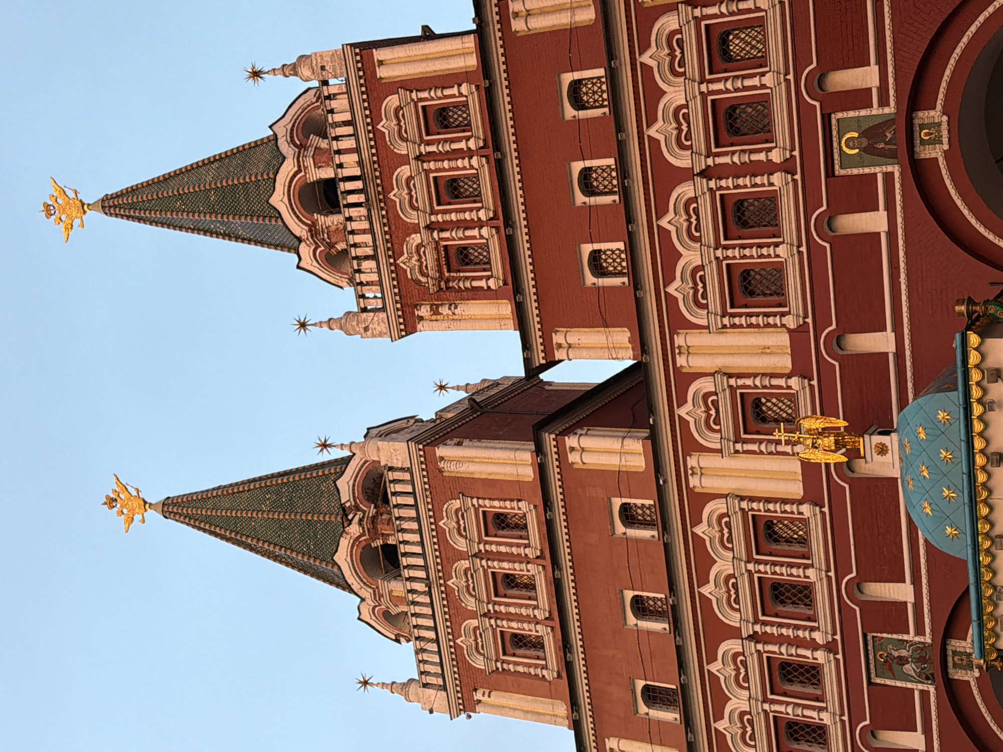 The image shows a close-up view of the upper sections of two towers of the State Historical Museum, located on Red Square in Moscow, Russia. The towers are constructed of red brick and feature ornate white detailing around the windows and decorative elements. Both towers are topped with green, conical roofs, each crowned with a golden double-headed eagle. The sky behind the towers is a clear, light blue. The towers are adorned with numerous small, square windows, many with intricate latticework. In the foreground, a portion of a blue dome decorated with golden stars is visible, along with a golden statue of an angel with outstretched wings holding a cross. There are also small mosaic images on the facade, including one depicting Saint George slaying a dragon.