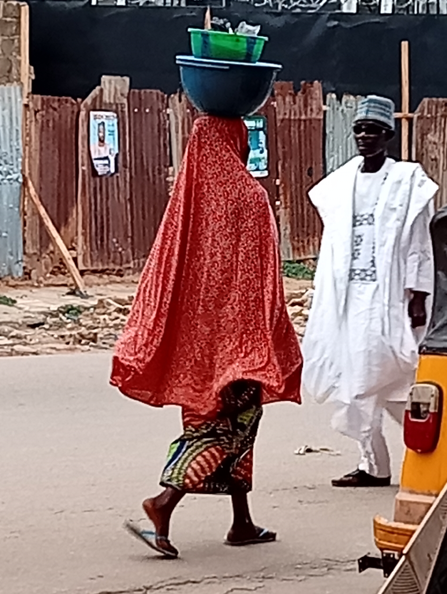 A woman is walking in the city of Maiburiji, Nigeria. She is carrying two basins on her head and is covered in a red cloth. She is wearing a colorful skirt and sandals. A man is walking behind her, wearing a white outfit, shoes and sunglasses. Behind them is a corrugated metal fence with a sign. A yellow vehicle is partially visible on the right side.