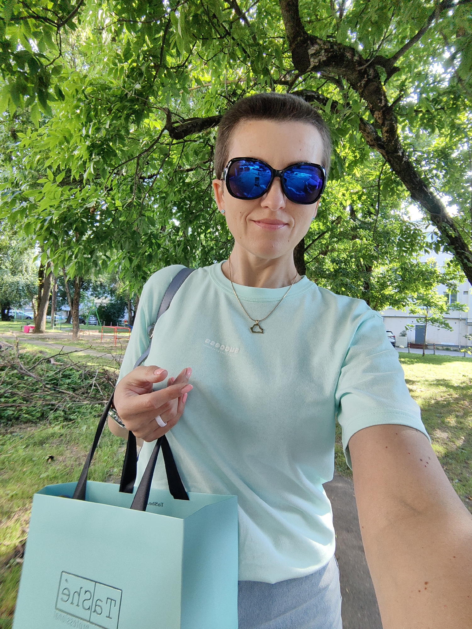 A woman with short dark hair and sunglasses is taking a selfie outdoors. She is wearing a light blue t-shirt, a necklace with a triangle pendant, and holding a light blue shopping bag. She is standing under a large tree with green leaves, and in the background, there are more trees, a building, and some playground equipment. The overall setting appears to be a park or a residential area. The image appears to be taken on a sunny day. The shopping bag has the logo "Tаgde" visible on it. Based on the visible text on the shopping bag, it is likely that the image was taken in Minsk, Belarus, as "Tаgde" is a Belarusian retailer.
