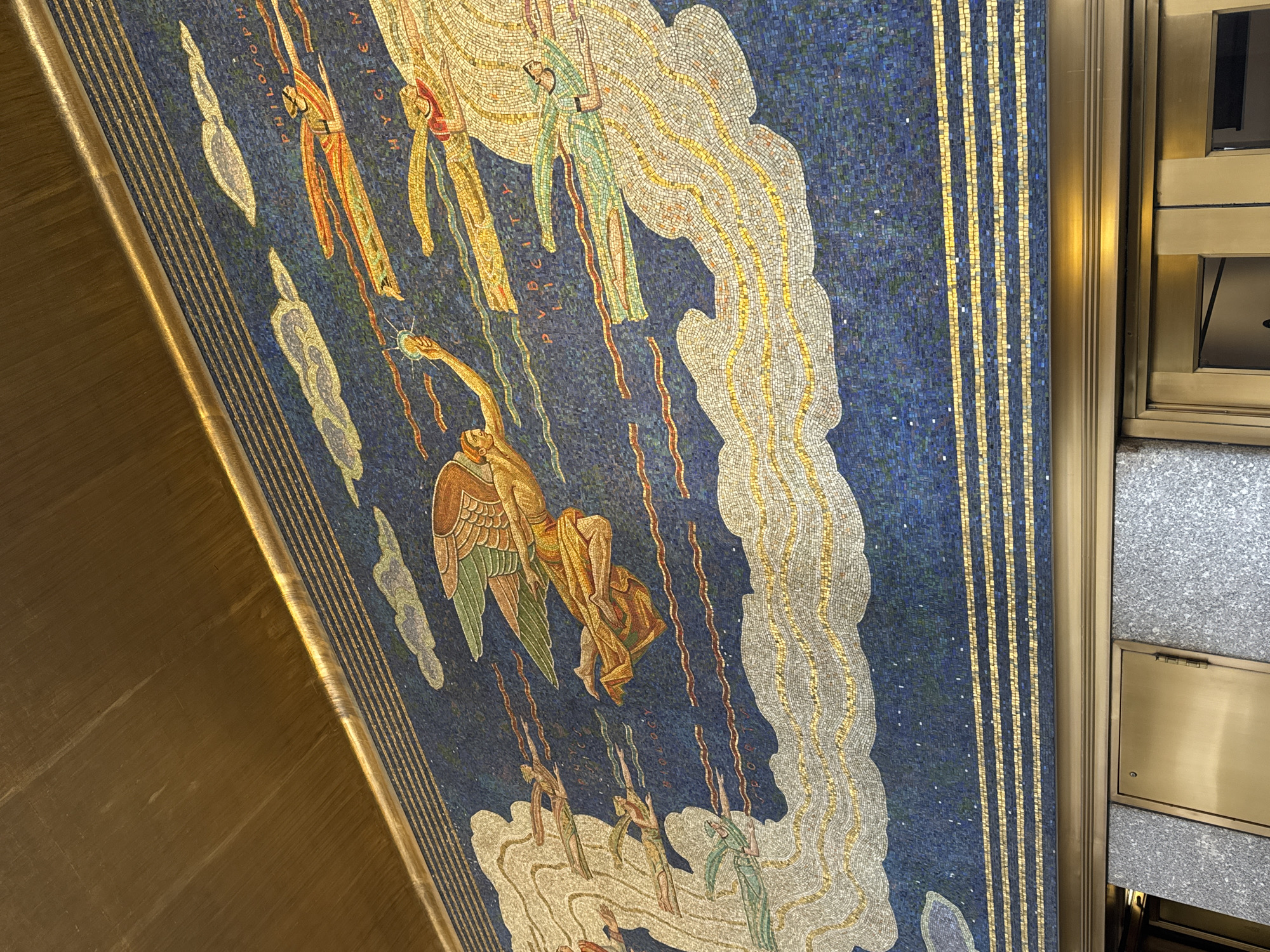 The image displays a large mosaic artwork titled 'The Spirit of Communications' (also known as 'Genius of Man') by Barry Faulkner, situated above the entrance to the Associated Press Building at 50 Rockefeller Plaza in Rockefeller Center, New York City, United States of America.
The mosaic features a deep blue, star-speckled background representing a sky, crossed by stylized white, grey, and gold clouds. Multiple human-like figures are depicted in flight. A central winged figure, with brown and green wings and a golden-orange robe, extends its left arm forward. To its left, an unwinged figure, garbed in orange and yellow, holds aloft a radiating, star-like object in its right hand. Smaller, less distinct figures, some trailing colorful streamers, are distributed throughout the mosaic, some partially integrated into cloud formations. Golden tesserae spell out partial words within the blue background, including 'PHILOSOPH,' 'HYGIEN,' 'PHYSI,' 'BIOLOGY,' and 'PUBLICITY.'
Above the mosaic is a wide panel of brushed golden metal, serving as a ceiling or architectural trim. Below the artwork, a golden frame separates it from a lower architectural section composed of speckled grey granite walls and golden-colored panels, including a door or service panel on the left and an entrance structure on the right.