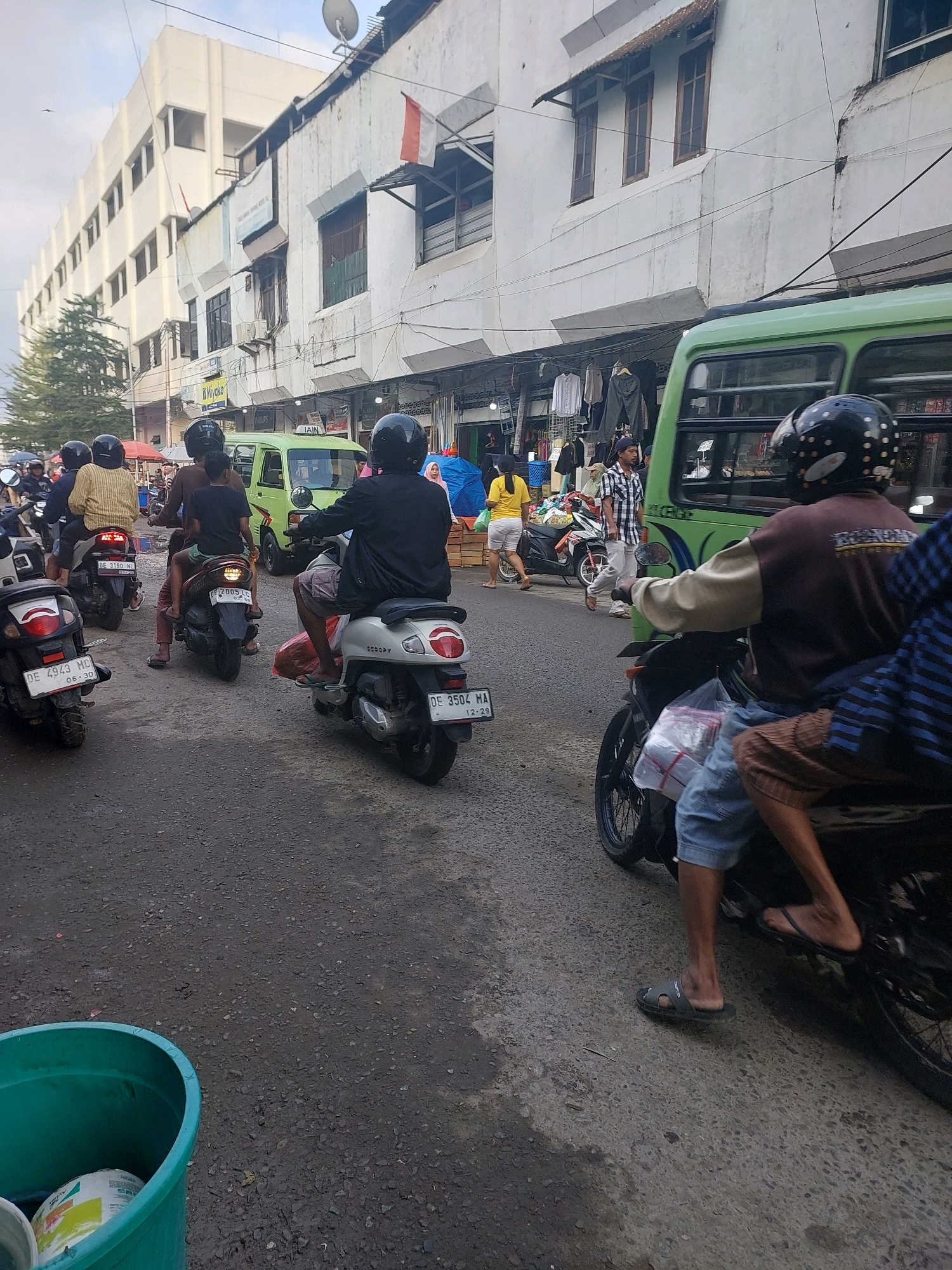 This is a ground-level, eye-level, medium shot of a busy street scene in Ambon, Indonesia, likely a market area, under an overcast sky. The foreground shows a section of asphalt road, wet and speckled with small stones. A large, green plastic trash bin is visible on the left, containing part of a cylindrical object with a label that includes yellow and white.
In the mid-ground, multiple motorcycles are either parked or moving, creating a traffic jam. One motorcycle in the foreground on the right has a rider wearing a brown and cream jacket, light blue denim shorts, and dark flip-flops. The motorcycle itself is dark-colored and has a plastic bag with items wrapped in white plastic tied to its front. Further left, a silver scooter is ridden by a person wearing a black jacket and plaid shorts, carrying a red bag on the back. Another scooter to the left of this one is ridden by a person in a brown shirt and dark pants. In the background, a green van is visible, along with other people and motorcycles.
The background features a multi-story building with a white facade. The lower level appears to be shops with clothing racks displaying garments, and awnings protruding over the sidewalk. The upper floors have numerous windows. A satellite dish is mounted on the roof of the building. An Indonesian flag is also visible, attached to the building. Trees can be seen to the left of the building. The overall impression is of a vibrant, active commercial street.