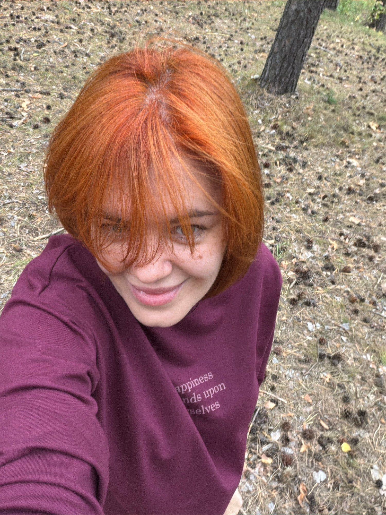 A young woman with short, bright orange hair is taking a selfie outdoors. She is wearing a dark purple long-sleeved shirt with white text that reads, "happiness depends upon ourselves". Her face is partially obscured by her bangs, but she is smiling and looking towards the camera. She has freckles on her cheeks and nose. The background consists of dry grass, fallen leaves, pinecones, and the base of a tree trunk, suggesting a park or forest area. The ground is uneven and covered with natural debris. The lighting suggests it is daytime.