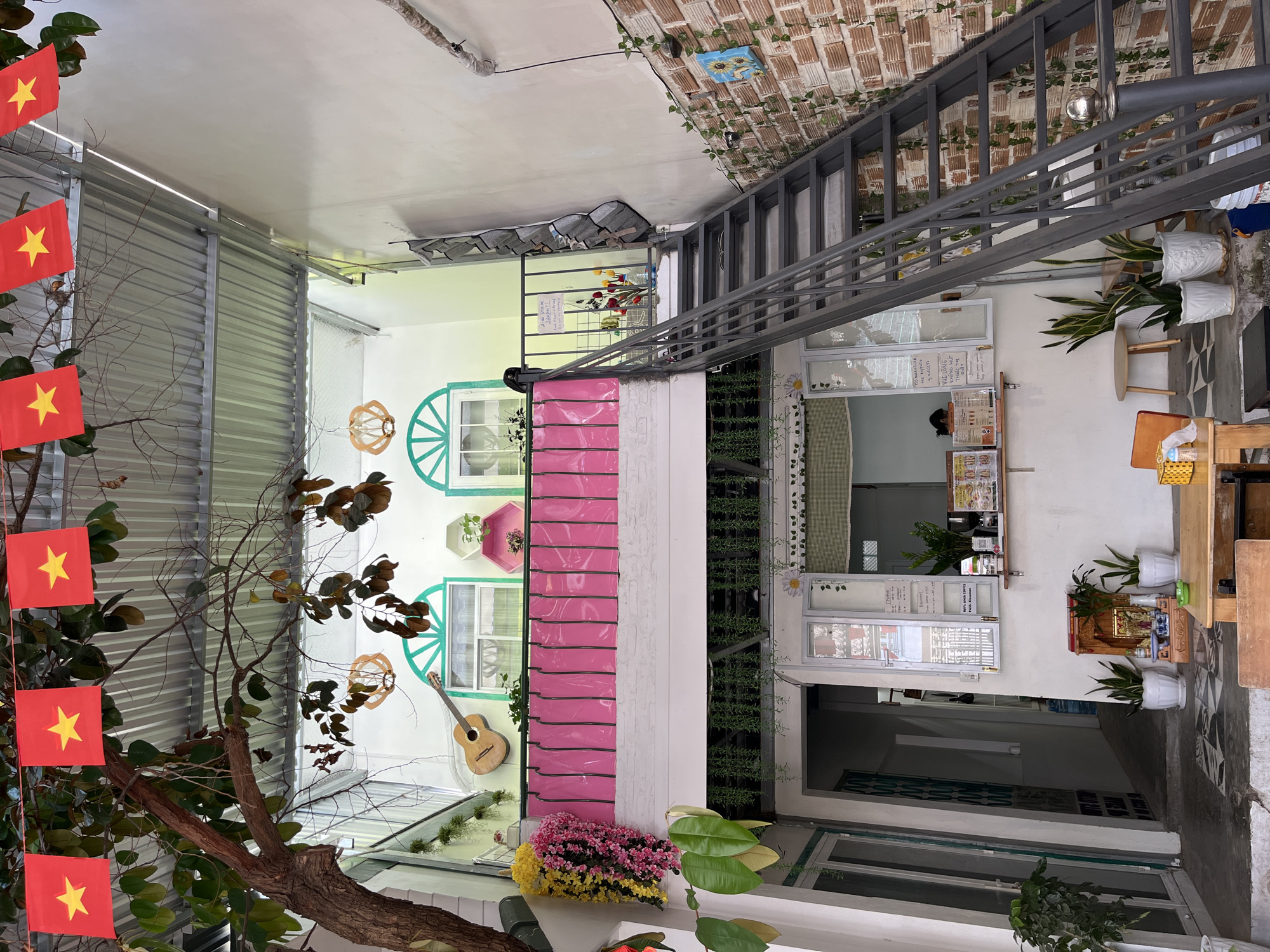 Here is a detailed description of the image:
**Scene:** The image captures a charming and colorful outdoor space, possibly a café or a courtyard, in what appears to be a residential or mixed-use area. The space is creatively decorated with a blend of traditional and contemporary elements.
**Architecture and Décor:**
- Walls are painted in a variety of colors, including white, pink, and light green. Some walls feature whimsical decorations like painted window frames and a guitar.
- There are visible brick elements and metal staircases, adding an industrial touch to the otherwise whimsical setting.
- Greenery is abundant, with plants in pots, hanging vines, and vertical gardens.
- Vietnamese flags are prominently displayed, indicating the location.
- A small counter area suggests food and drink preparation.
**Objects:**
- Tables and chairs are set up for customers.
- Various decorations, including flowers, wall art, and plants.
- A guitar is mounted on the wall.
- Vietnamese flags are attached to a tree.
- Stairs on the right side.
**People:**
- A person can be seen behind the counter.
**Environment:**
- The lighting indicates daylight, suggesting an outdoor or semi-outdoor setting.
- The presence of plants and natural elements indicates a courtyard or garden-like atmosphere.
**Location:** The presence of Vietnamese flags indicates the setting is in Vietnam.