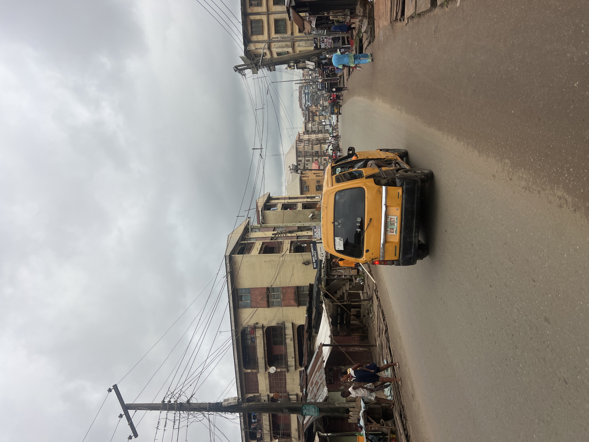 This is a medium-angle, eye-level shot looking down a dusty, asphalt road in Nkpor, Nigeria. The overcast sky suggests it might be a cloudy day, or possibly evening. The street is lined with multi-story buildings, some appearing weathered and possibly residential or commercial. There are many utility poles with a dense network of wires crisscrossing overhead. Along the sides of the road, there are makeshift structures, possibly market stalls or shops, with corrugated metal roofs and wooden supports.
In the foreground, a yellow minibike-taxi (known locally as an "Okada" or "Keke Napep") is driving away from the camera. The license plate reads "NSK 913ZY". To the left of the road, three individuals are walking, two young men and a woman in a dark dress. To the right, further down the road, a person in a light blue traditional outfit is walking away from the camera. More people are visible in the distance, some walking and some gathered near buildings. A white car is also visible further down the street. The general impression is of a busy, somewhat informal urban environment.