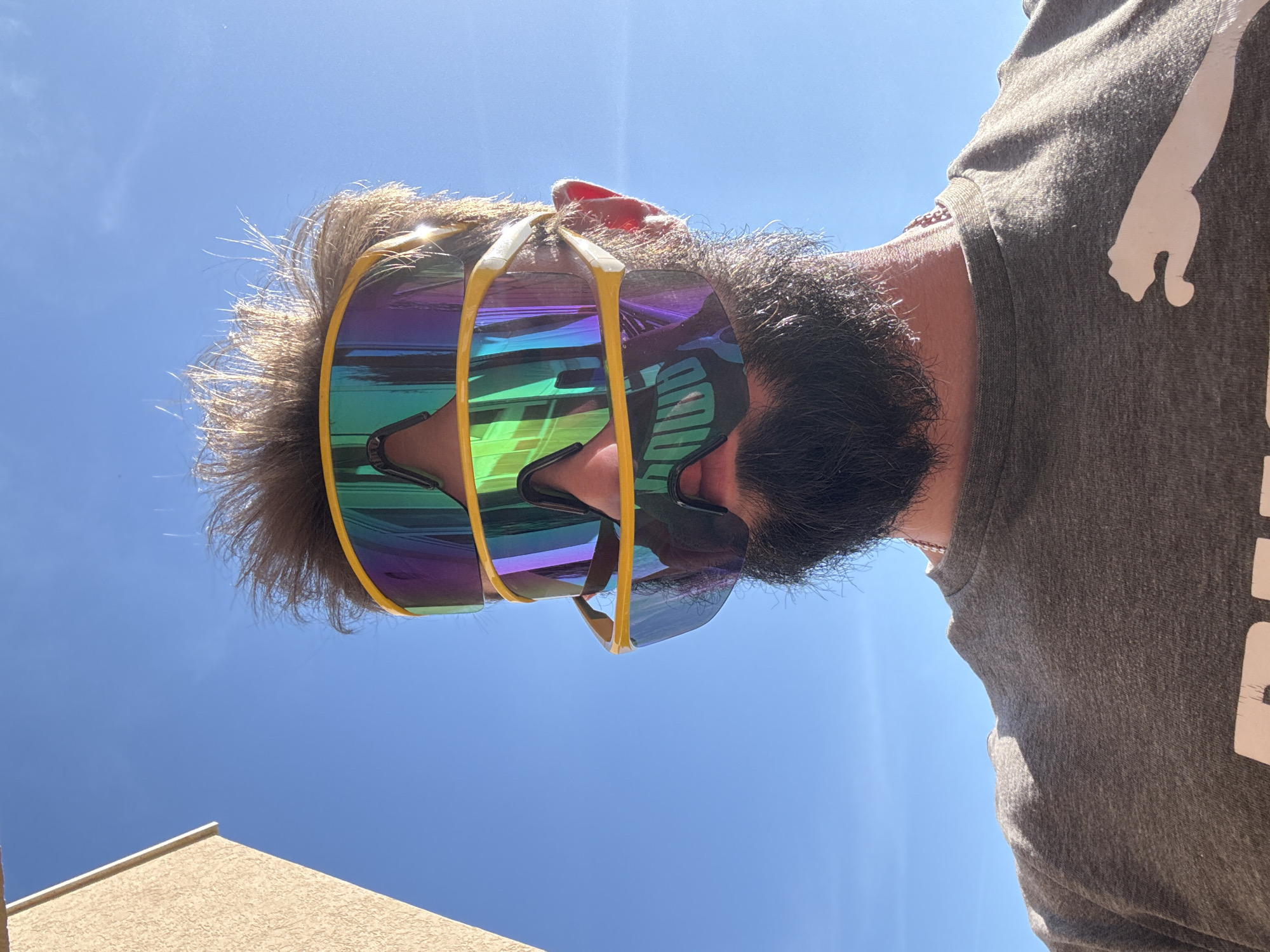 A man with a beard and wearing sunglasses stands in Šušanj, Montenegro, looking up at the sky. He is wearing a grey t-shirt with a white Puma logo on the right shoulder. The sunglasses have yellow frames and reflective lenses with green, blue, and purple tints. The sky is clear and blue. The top of a tan-colored building is visible in the lower left corner of the frame.