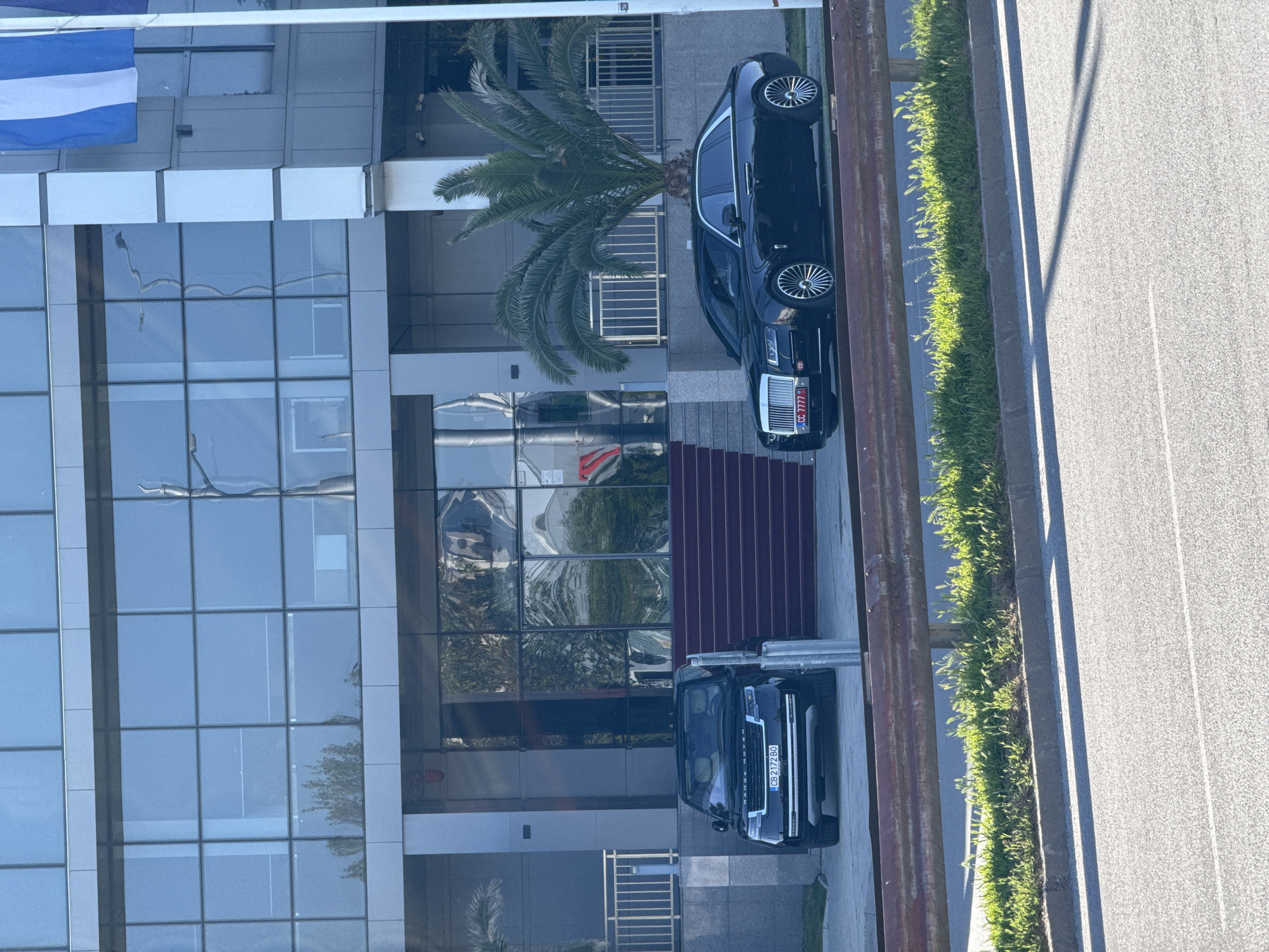 The image captures a modern, multi-story building featuring extensive glass paneling and grey architectural elements. In front of the building, two luxury black vehicles are parked. A black Rolls-Royce is positioned to the right, displaying the license plate "CC 7777". Closer to the building on the left, a black Range Rover is parked, bearing the license plate "CB 2172 BO".
Between the parked cars and the building's facade, a tall palm tree with lush green fronds adds a tropical touch. A set of red carpeted steps leads up to what appears to be an entrance. To the far left, a flag with horizontal blue and white stripes is visible hanging from an upper section of the building.
The scene is brightly lit, indicative of daytime with clear, sunny weather, as evidenced by strong shadows cast on the asphalt road and reflections on the glass. A strip of vibrant green grass separates the building's immediate vicinity from the wide asphalt road, which features clear white lane markings. The overall setting suggests a modern urban or corporate environment.
