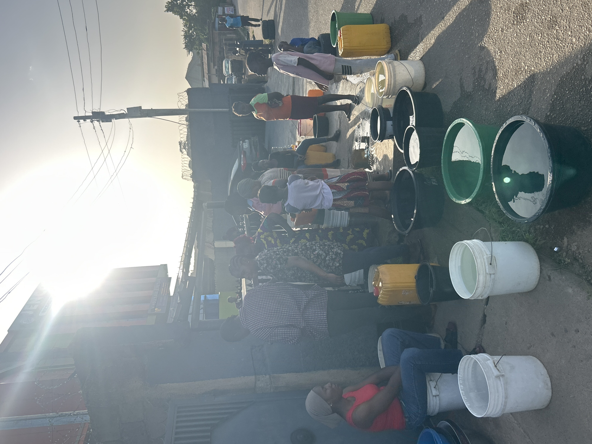 The image depicts a group of people in Jos, Nigeria, gathered along a roadside with various containers, including buckets and jerrycans. Many containers are filled with water. Some individuals are standing and appearing to fill or wait to fill their containers, while others are seated nearby. The setting suggests a water distribution point or a location where water is being collected. The background includes buildings, power lines, and other signs of urban infrastructure. The bright sunlight indicates it is daytime.