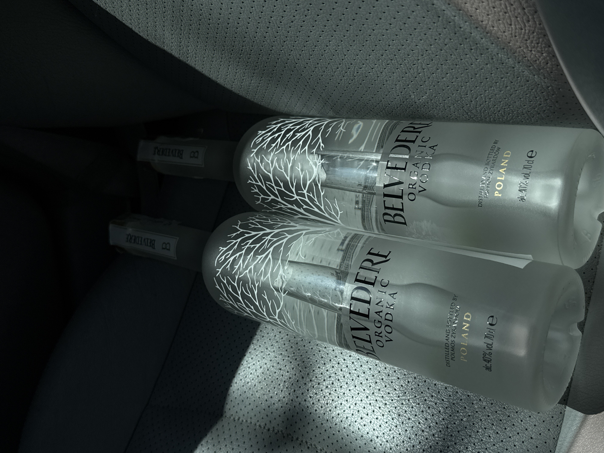 Two frosted glass bottles of Belvedere Organic Vodka are placed on a dark gray, perforated leather surface, likely the interior of a car. The bottles are partially obscured by sunlight, creating dappled shadows on the surface. Both bottles feature the distinctive Belvedere branding with a white tree silhouette and the text "BELVEDERE", "ORGANIC", and "VODKA". Below this, the text "DISTILLED AND BOTTLED BY POLMOS ZYRARDÓW", "POLAND", and "alc. 40% vol. 70 cl e" is visible. The scene is set within a vehicle, with the textured leather indicating it's likely seating or paneling. The overall lighting suggests it is daytime. There is no indication of the city of Tsarevo, Bulgaria.
