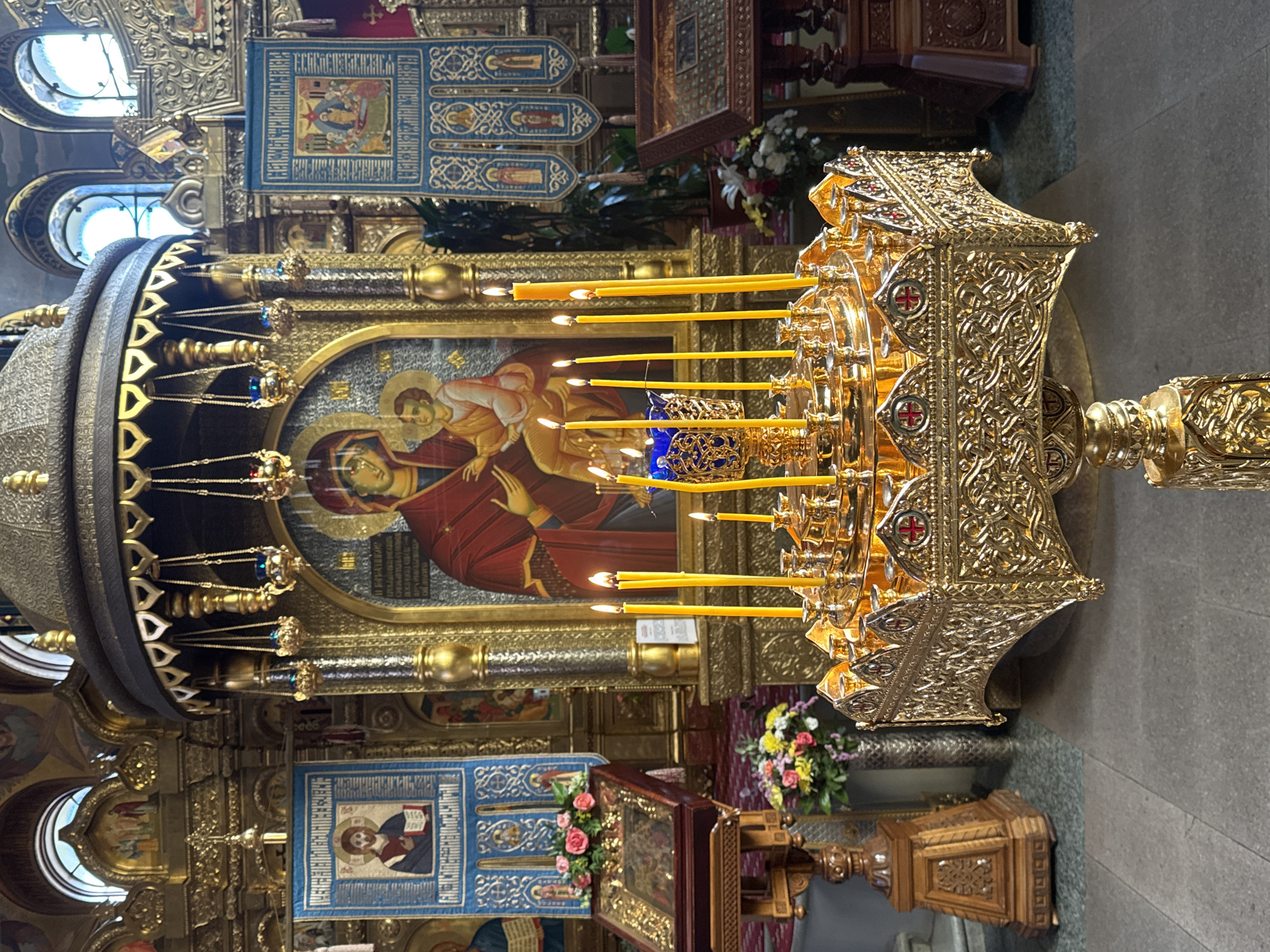 The image captures the interior of a Russian Orthodox Church, likely in Saint Petersburg, Russia, given the architectural style and religious iconography. A gold-colored, ornate candelabra stands in the foreground, holding numerous lit yellow candles. Behind the candelabra, a large icon depicts a religious scene, possibly the Virgin Mary with the Christ Child. Further back, additional icons and religious banners adorn the walls. The environment appears to be dimly lit, with the candles and gold accents providing the primary sources of light. Floral arrangements are visible in the background. The image is taken at eye level, possibly from a standing position within the church.