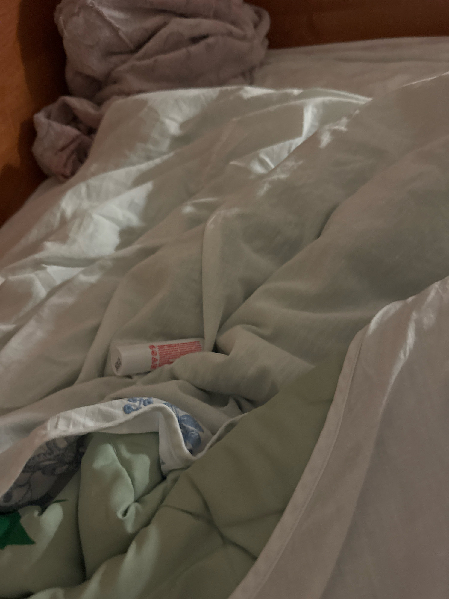 An unmade bed is shown with white and light green bedding. A small white tube with red writing, possibly a lip balm or medication, is partially visible under the white blanket. The scene is dimly lit, suggesting it might be nighttime or an indoor setting with minimal light. The overall impression is one of casual disarray. The location in Zavolzhye, Russia, is not depicted in the image.