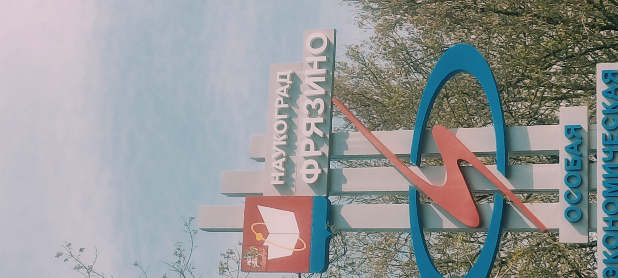 A sign in Fryazino, Russia, displays the city name in Cyrillic: "НАУКОГРАД ФРЯЗИНО". Below the text is a red square with a blue border containing a white book graphic surrounded by a gold atom model and a small emblem at the bottom. Beside this is a blue oval intersecting a red stylized 'M'. Beside this is the Cyrillic text "ОСОБАЯ ЭКОНОМИЧЕСКАЯ". The sign is located outside amidst foliage and a partially cloudy sky.