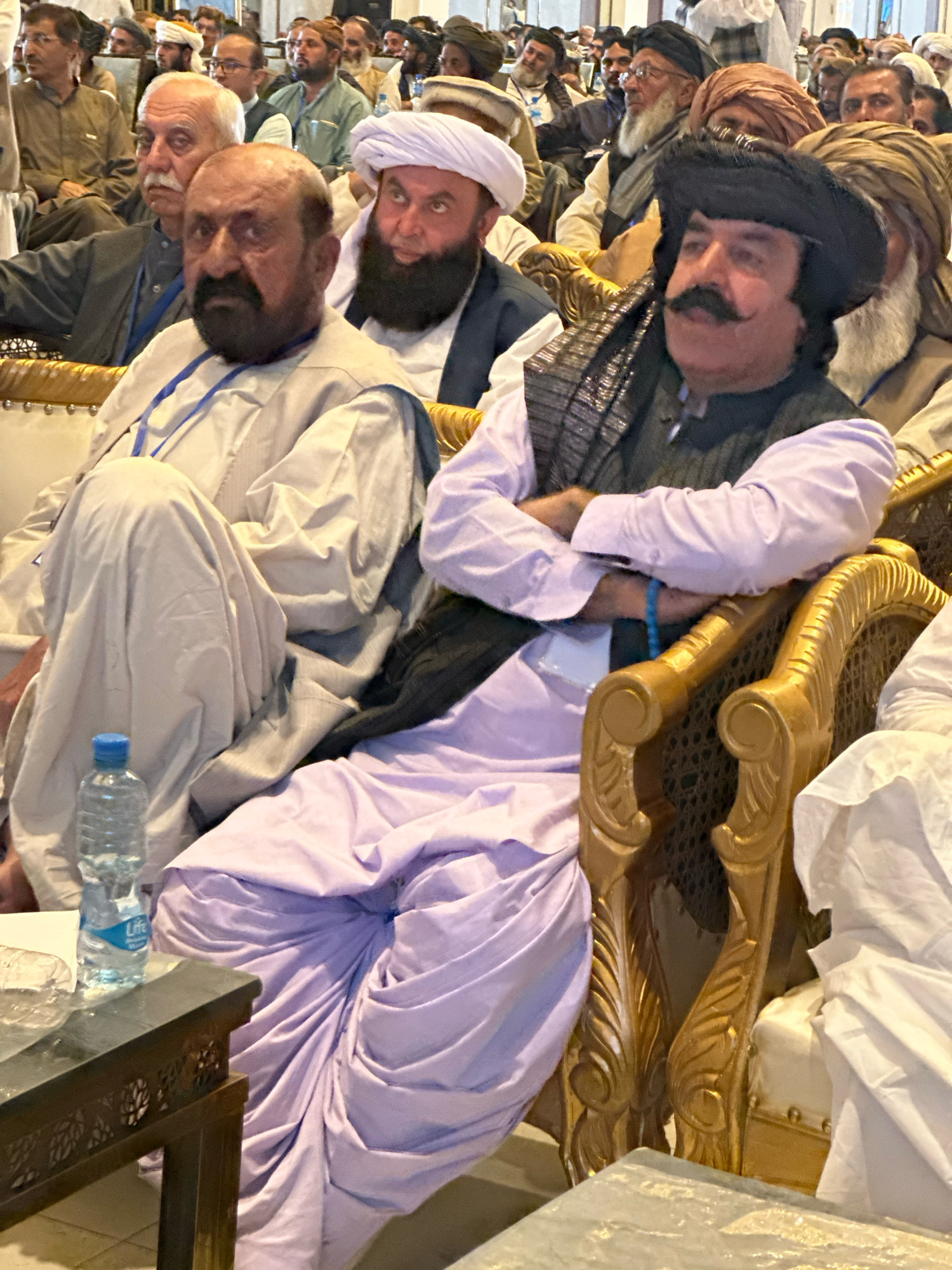 The image depicts a gathering of men in Quetta City Tehsil, Pakistan, likely an indoor event given the seating arrangements. In the foreground, two men are prominently seated on ornate golden chairs. The man on the left, with a prominent mustache and wearing a black turban and a dark vest over a light-colored shirt, is facing forward and appears to be speaking or being addressed. He has a blue bracelet on his left wrist. To his right, another man, also seated, is partially visible. He has a beard and is wearing a light-colored garment. A bottle of water and a plastic wrapper are on a small wooden table to the left of the frame. The background is filled with other attendees, many wearing traditional turbans and clothing, suggesting a cultural or political assembly. The overall atmosphere appears formal and possibly ceremonial.