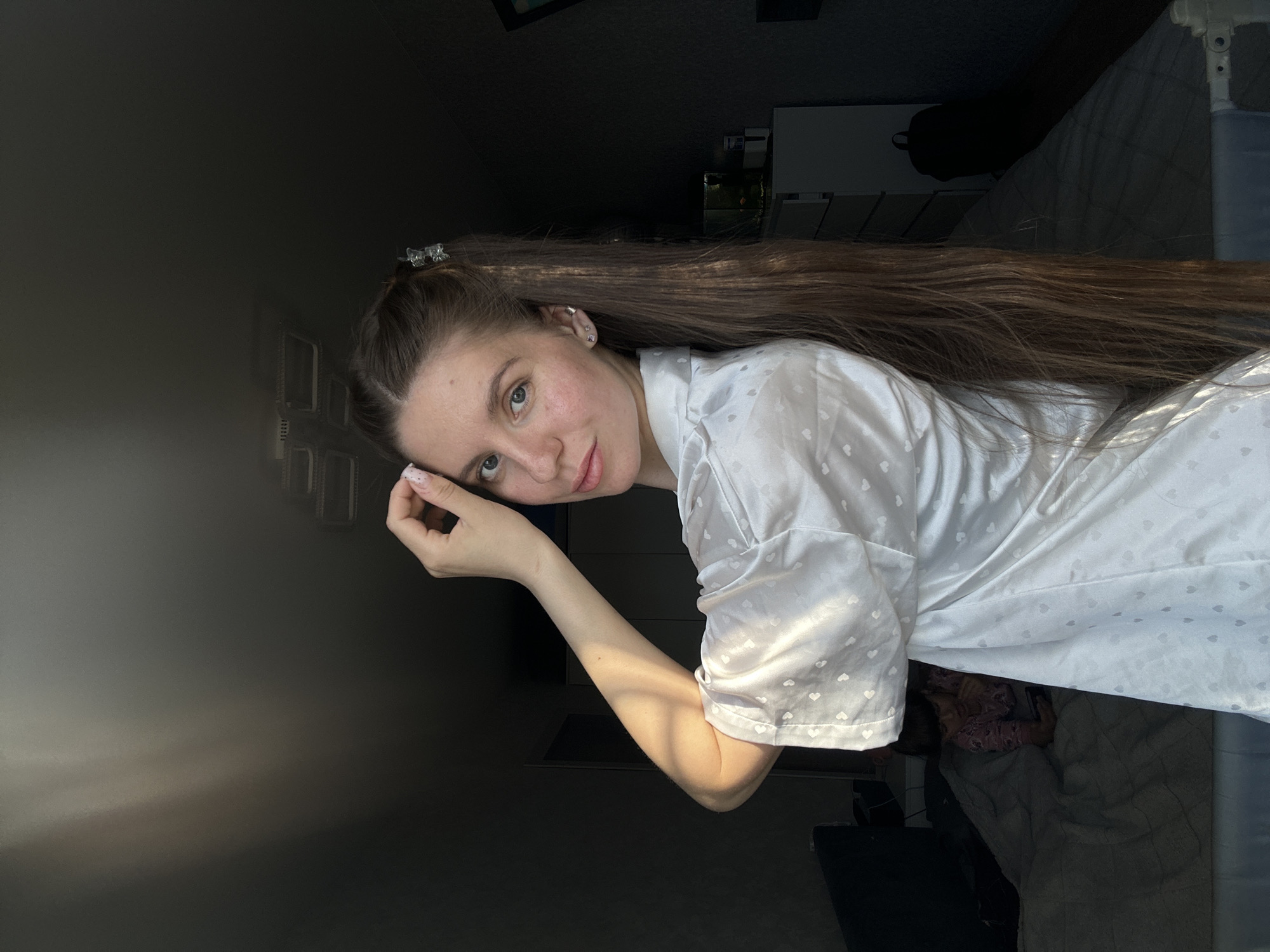 A young woman with long brown hair and blue eyes is posing indoors. She is wearing a white robe with small heart patterns. She has a silver hair clip in her hair. The background is dark and indistinct, but a wall with window panels can be seen. The location of Klin, Russia, is not evident from the image.