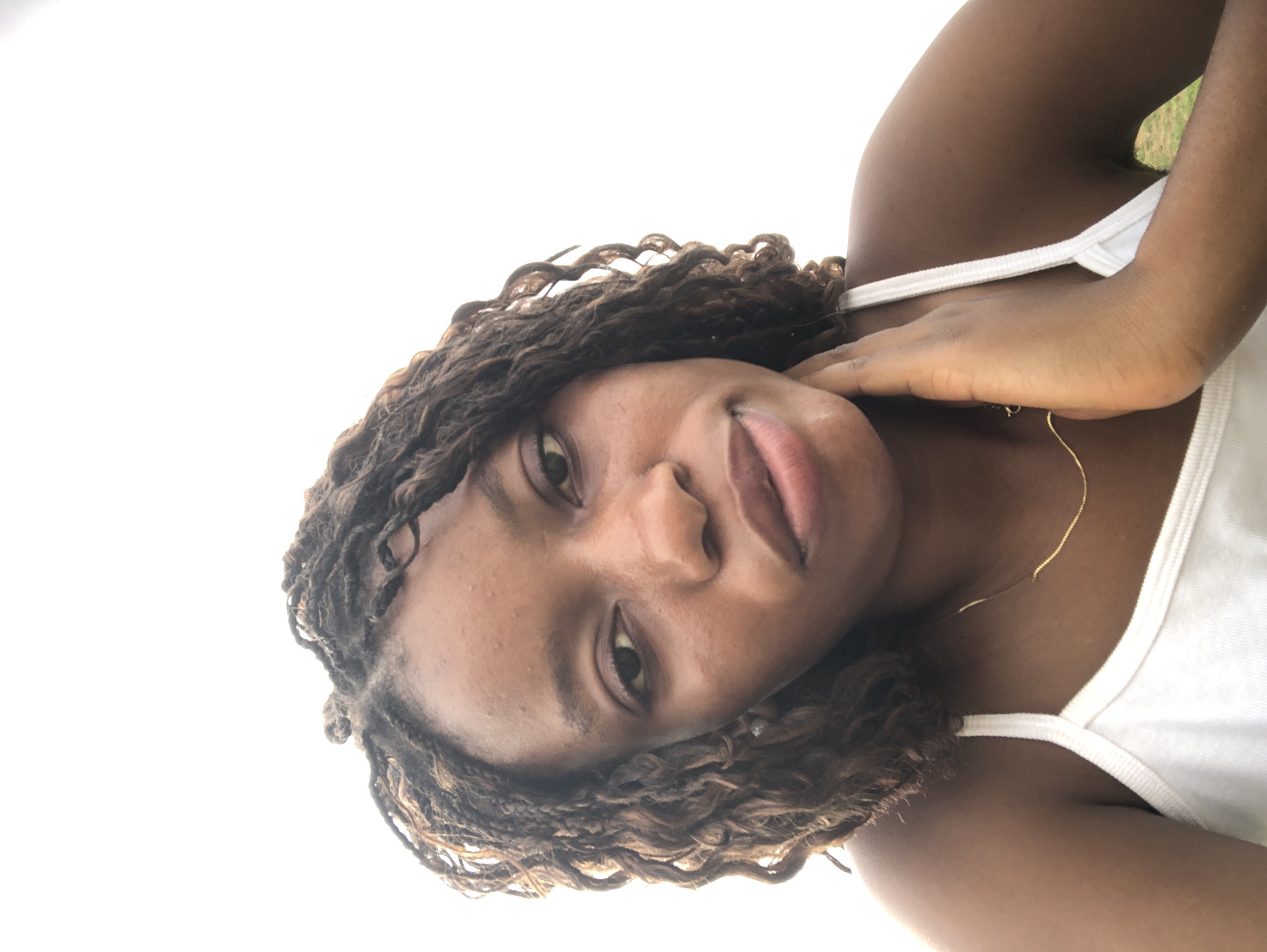 A close-up, rotated portrait of a young, dark-skinned woman is positioned against a bright, sky-like background, suggestive of an outdoor setting in Kashere, Nigeria. The woman's face, predominantly featuring her eyes, nose, and lips, is in sharp focus. Her hair is styled in twists or braids with curly ends. She wears a white tank top. Her right hand is gently placed on her neck, and a thin gold necklace is visible around her neck. The light suggests it is daytime.