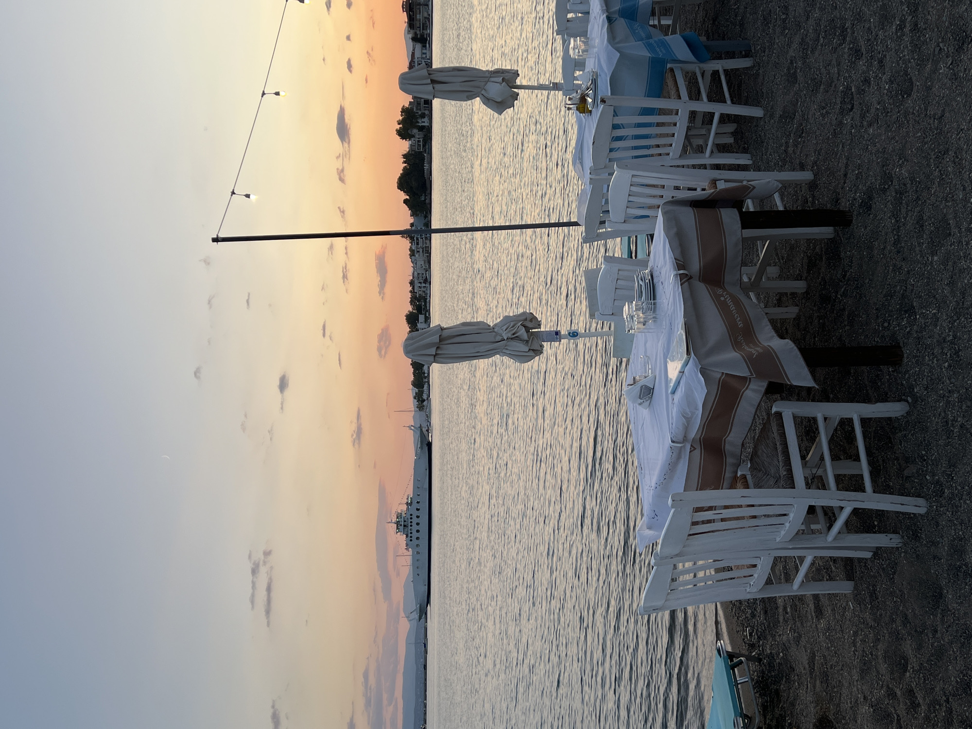 An outdoor dining scene is captured at sunset in Eretria, Greece. In the foreground, several white wooden chairs surround tables draped with white tablecloths, some adorned with a decorative brown trim. Plates, glasses, and bottles of condiments are set on the tables, ready for diners. Closed white umbrellas stand behind the tables, providing shade. The ground beneath the tables and chairs is sandy and pebbly.
Beyond the dining area, a calm body of water stretches to the horizon. A large white ferry boat is prominently featured sailing on the water in the mid-ground. In the background, the coastline of Eretria is visible, with a scattering of buildings and trees silhouetted against the vibrant sunset sky. The sky displays a gradient of colors, transitioning from pale blue at the top to soft oranges and pinks near the horizon, with a faint crescent moon visible in the upper left portion of the image. String lights are suspended from a pole above the tables, adding to the ambiance of the setting. The overall scene depicts a tranquil evening by the sea, with the promise of a meal outdoors.
