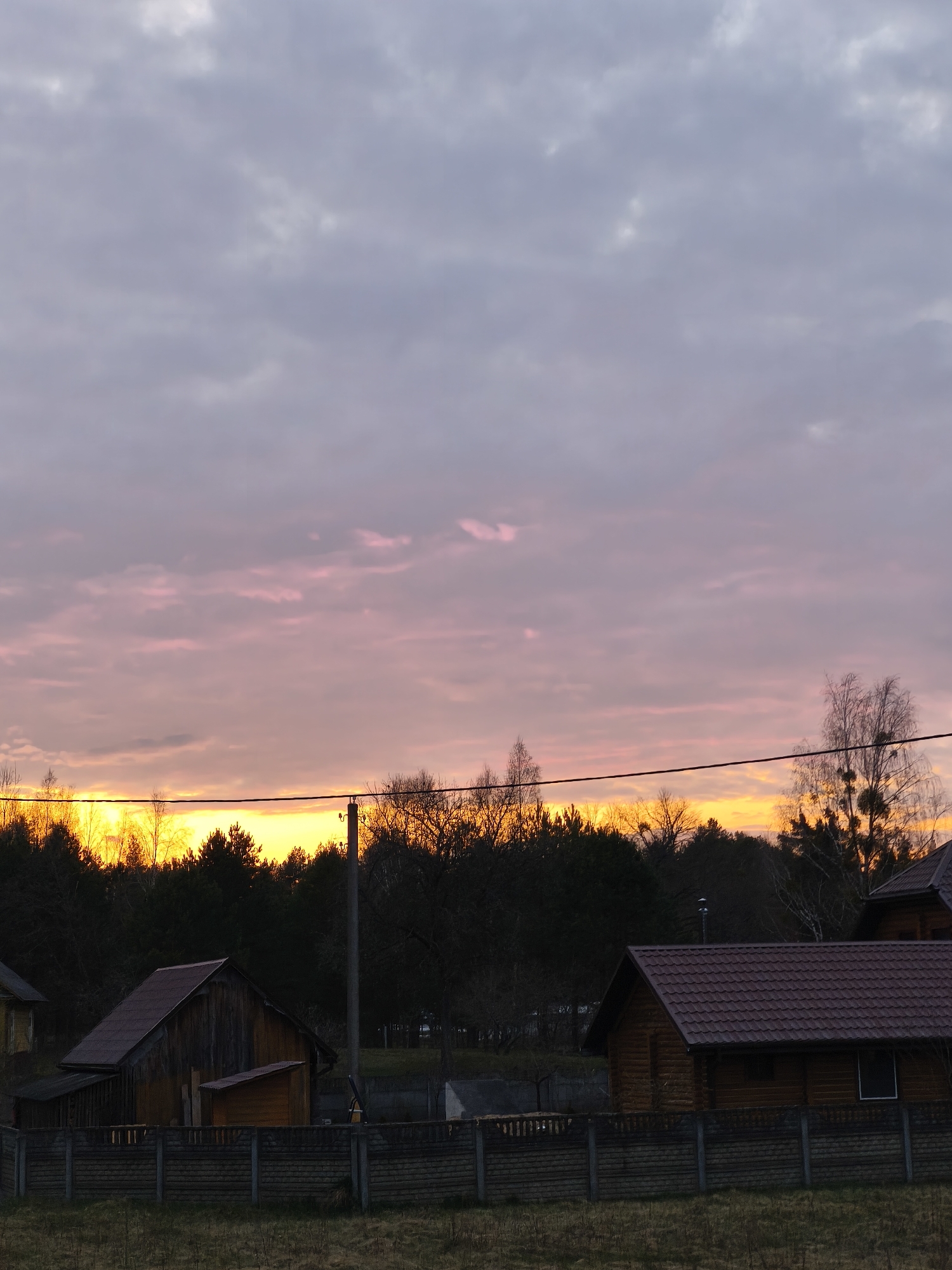 A photograph shows a landscape at dusk, possibly in Парэчча, Belarus. In the foreground, there is a grassy field, followed by a low concrete and wooden fence. Behind the fence are two small wooden buildings with red-tiled roofs. A power line stretches horizontally across the scene. In the midground, there is a dense line of trees, with a vibrant orange glow appearing behind them, suggesting a setting sun. The sky above the trees transitions from orange near the horizon to pink and purple towards the top of the frame, indicating the fading daylight.