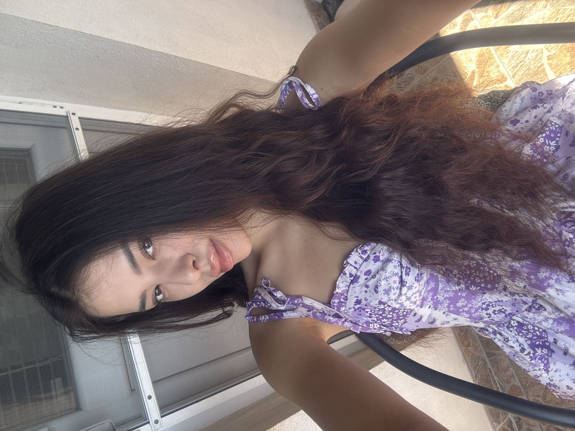 A young woman is seen from an elevated, angled perspective, possibly a selfie. Her long, wavy brown hair dominates the upper half of the frame. She has fair skin and appears to be wearing lip gloss.
She is wearing a floral-patterned dress with purple and white hues. Part of a black chair is visible, suggesting she is seated.
The background consists of a window with a screen, a beige wall, and tiled flooring, suggesting an outdoor or balcony setting.
Given the request to identify the location in Plovdiv, Bulgaria, and the limited visual clues, it's impossible to pinpoint the exact location based solely on the image. However, the architecture and overall aesthetic could be consistent with residential areas in Plovdiv.