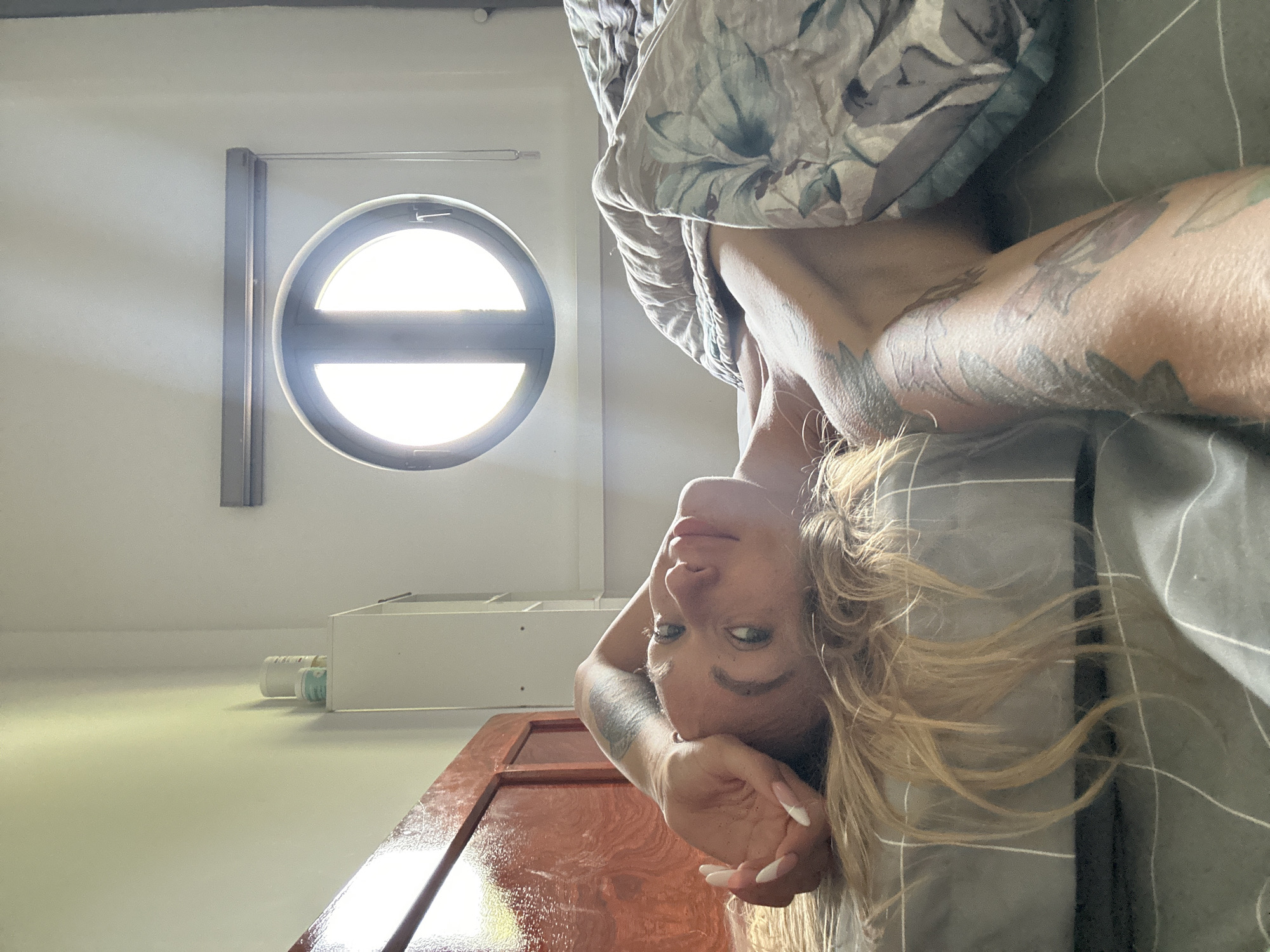 A woman lies in bed, her head propped on her arm. She is looking up and to the left, her expression relaxed. Her blond hair is spread around her head on the pillow. She has tattoos on her arm and shoulder. The room is lit by sunlight coming through a round window with a dark frame. A floral comforter is pulled up to her chest. A wooden headboard is visible to the left of the frame. A white cabinet with two jars on top is in the background to the left, and a white wall with a subtle gradient of light and shadow is on the right.
