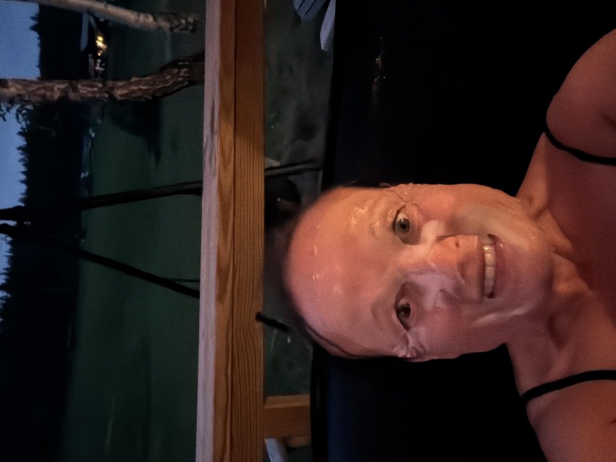The image shows a close-up of a person wearing a clear sheet mask, with their eyes and mouth visible. The person is looking directly at the camera and smiling. They appear to be in a dimly lit outdoor setting, with a wooden railing in the foreground and a dark, wooded landscape in the background. Lights are visible in the distance, suggesting a populated area. The overall scene suggests a nighttime setting, possibly in the city of Kongsberg, Norway, given the context of the prompt, though no specific landmarks confirm this location. The image focuses on the face of the person, with the environment serving as a muted backdrop.
